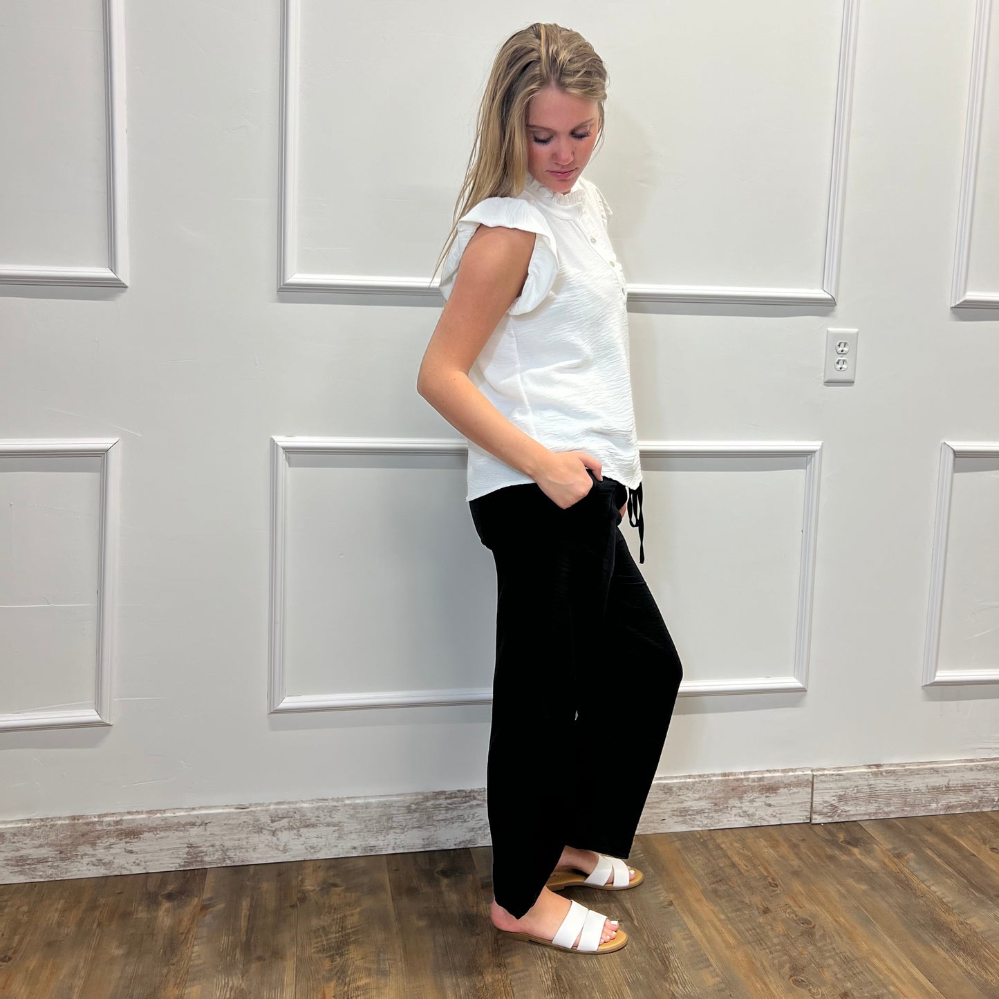Woman wearing a white blouse and black pants standing in a room with white paneled walls.
