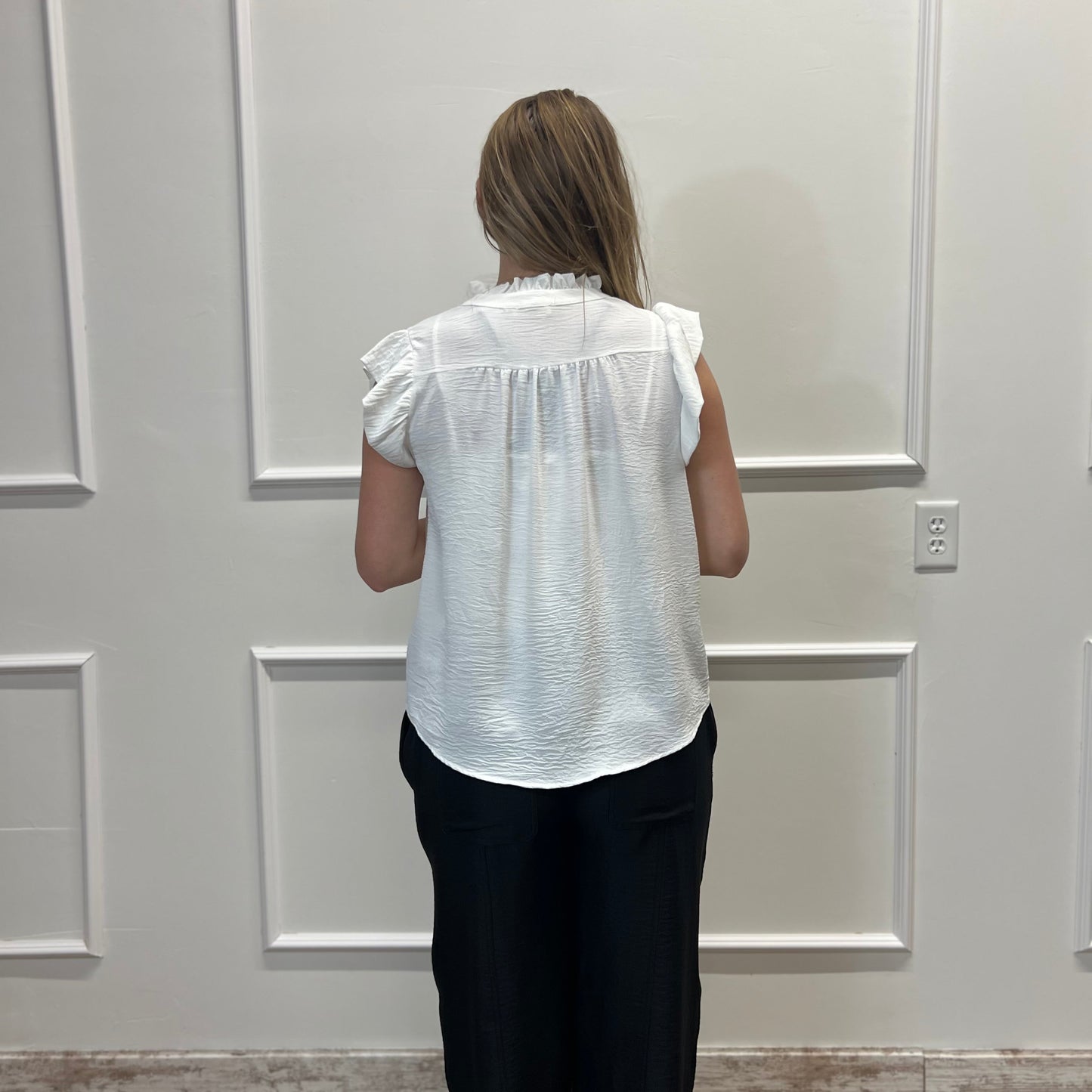 Person wearing a white blouse and black pants standing in front of a white paneled wall.