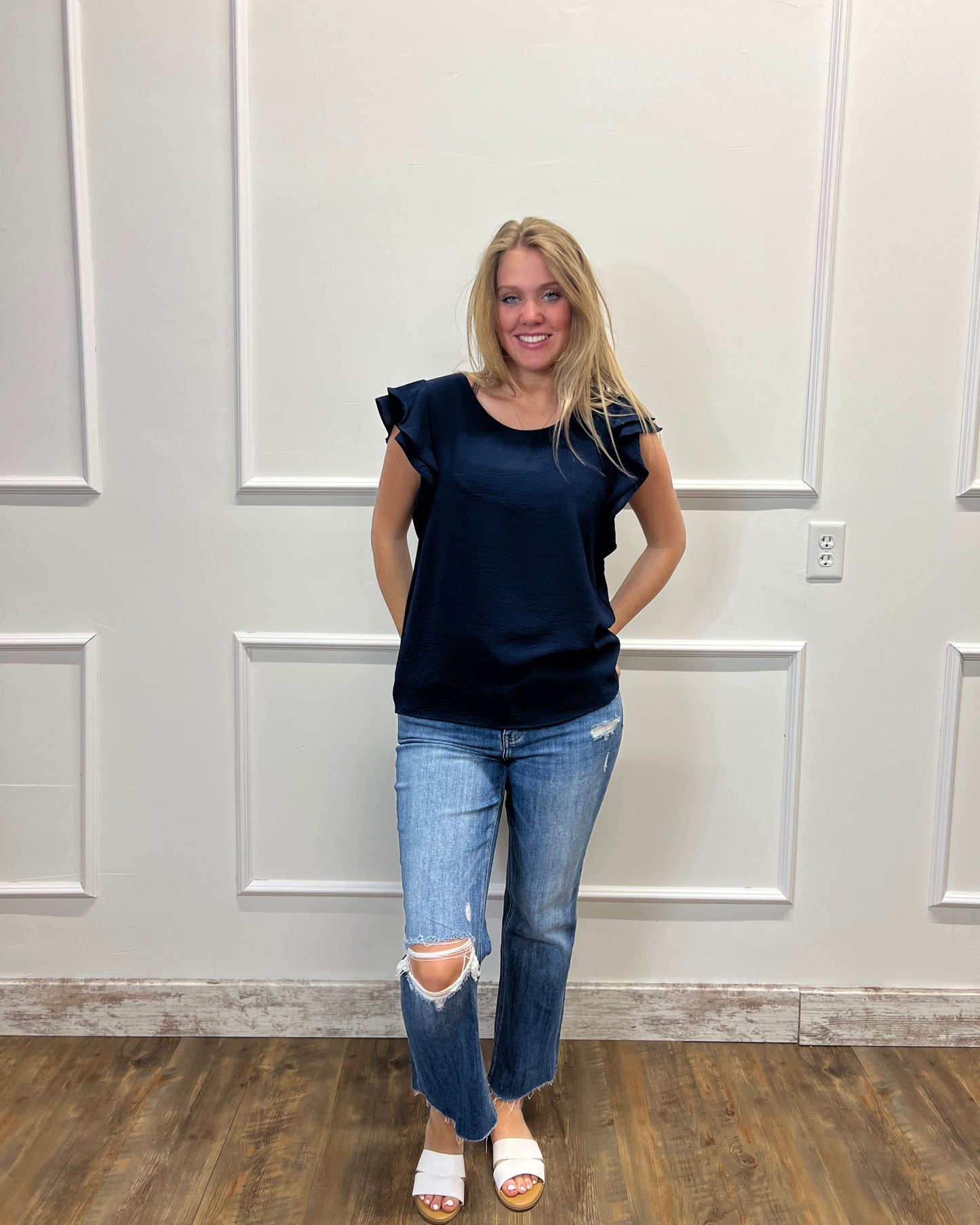Woman wearing a navy blue top and jeans standing against a white paneled wall.