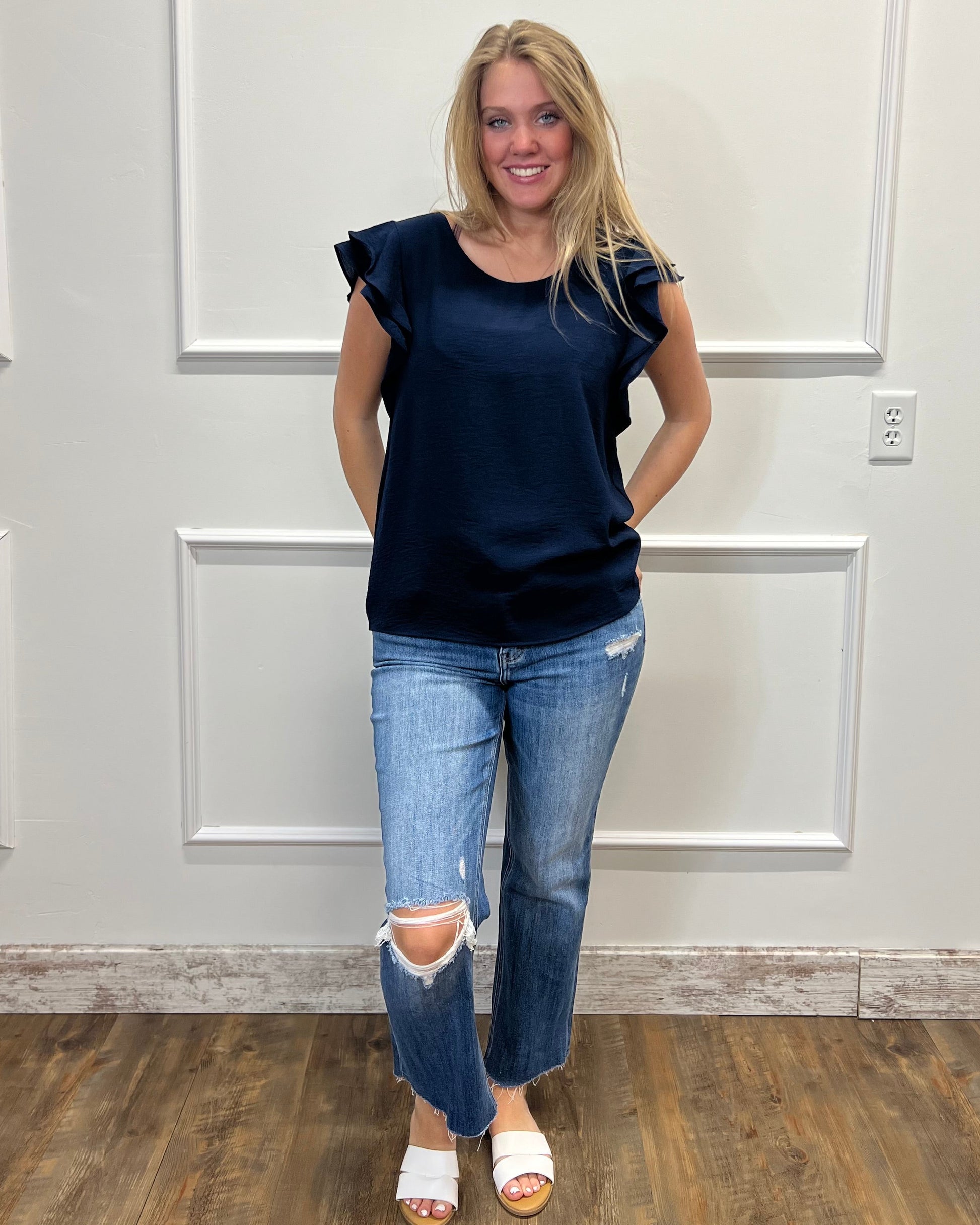 Woman wearing a navy blue top and ripped jeans standing against a white paneled wall.