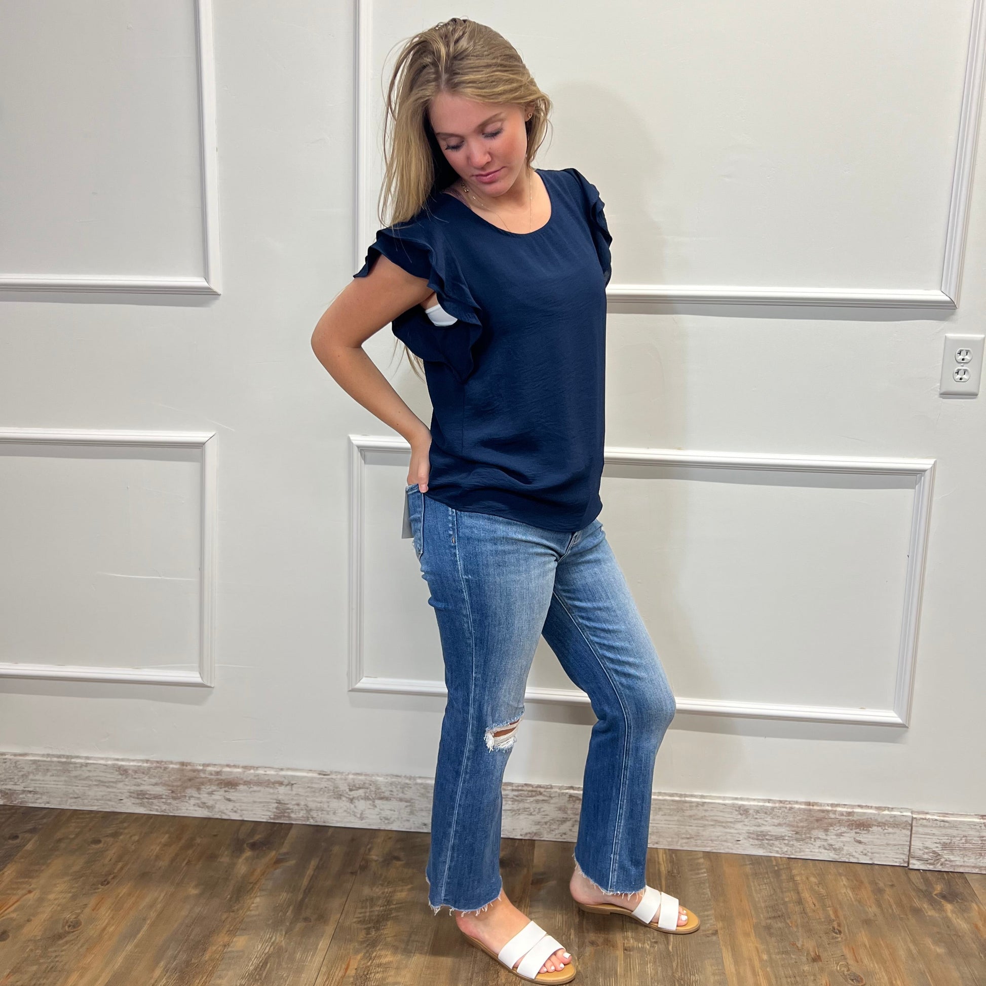 Woman wearing a navy blue top and jeans standing against a white paneled wall.