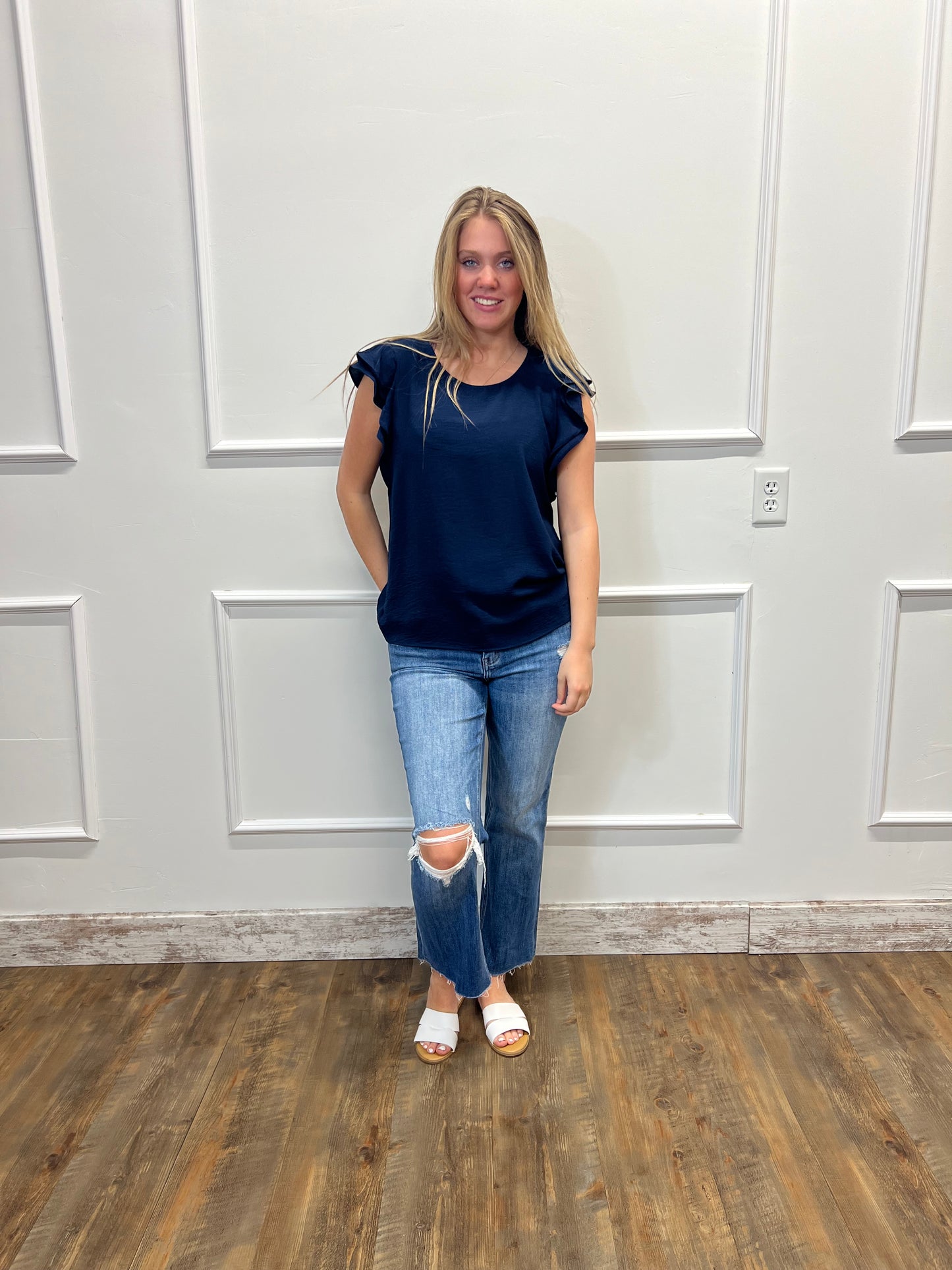 Person wearing a blue top and jeans standing in a room with white walls and wooden flooring.