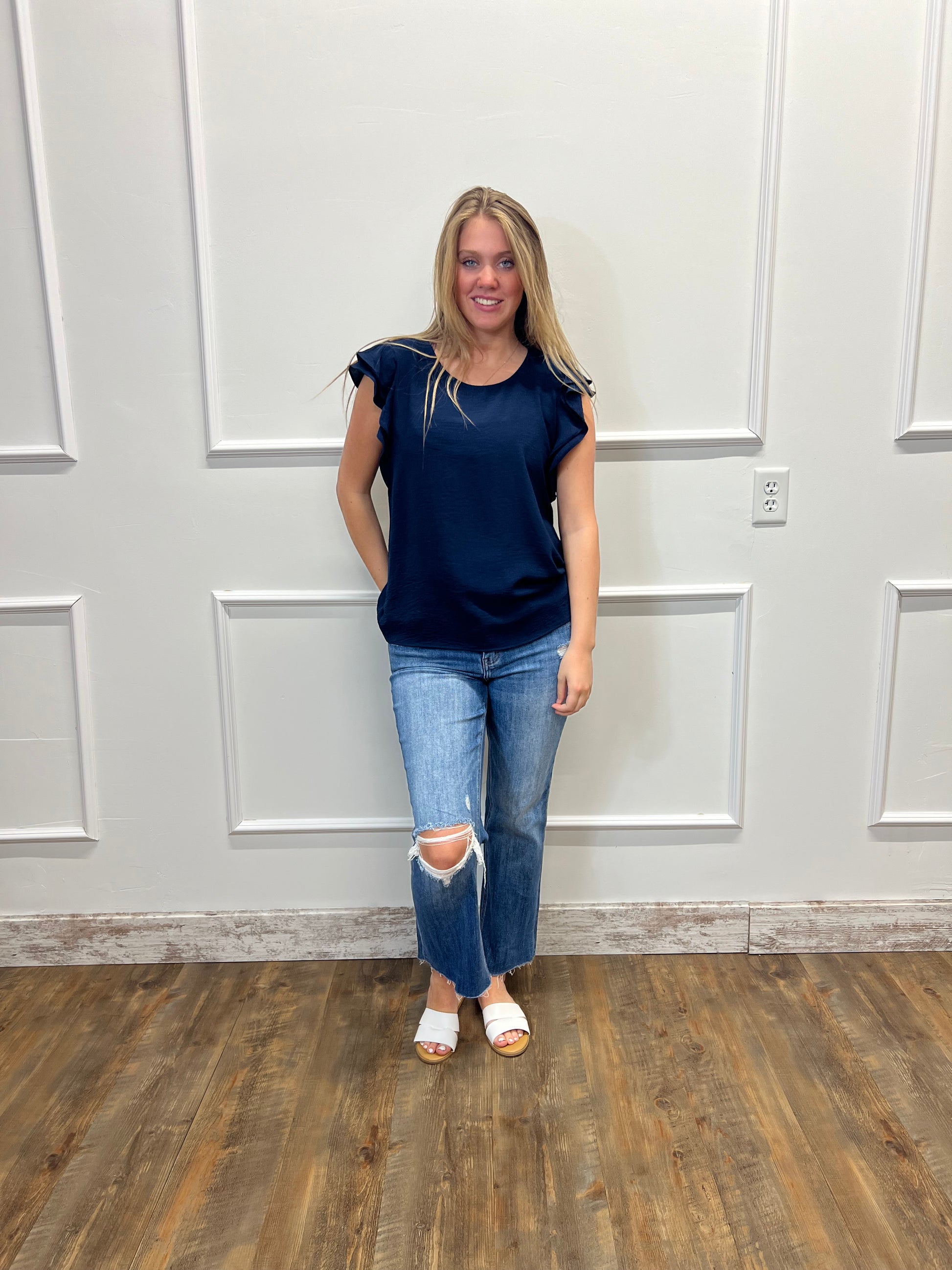 Person wearing a blue top and jeans standing in a room with white walls and wooden flooring.