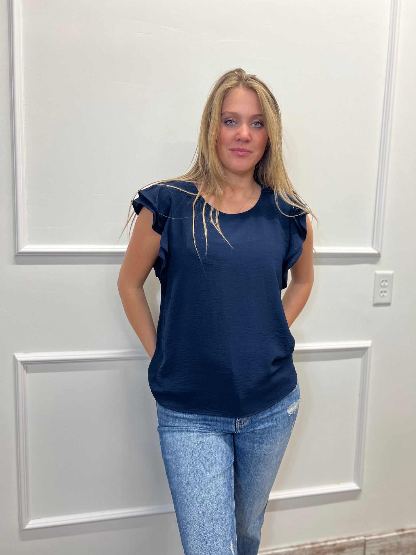 Woman wearing a navy blue top and jeans standing against a white paneled wall.