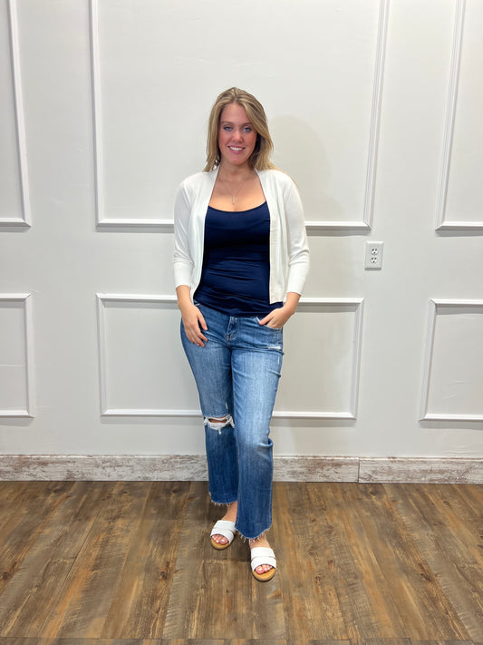 Woman wearing a navy top, white cardigan, blue jeans, and white sandals standing against a white paneled wall.