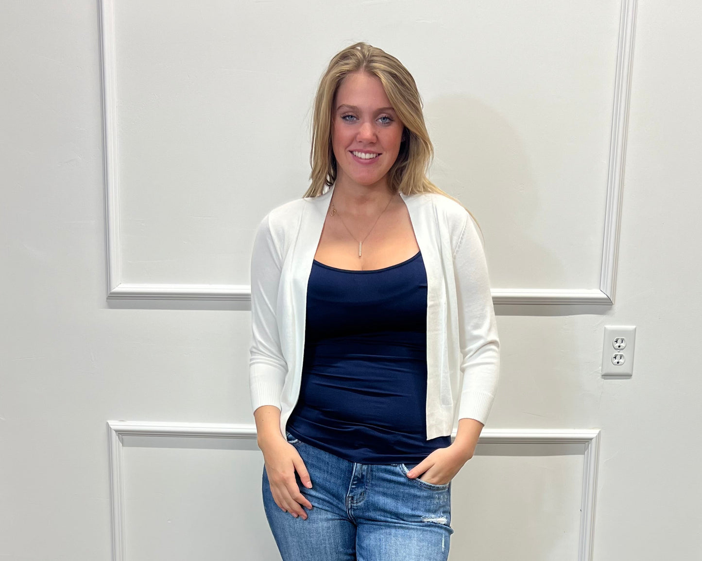 Woman wearing a navy top, white cardigan, blue jeans, and white sandals standing against a white paneled wall.