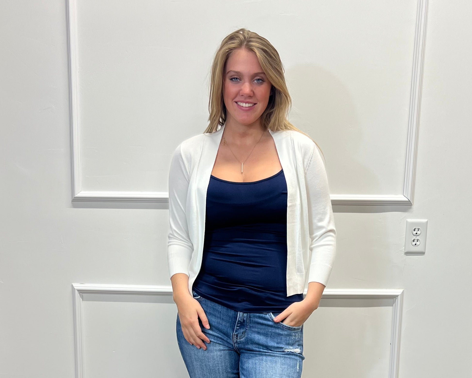 Woman wearing a navy top, white cardigan, blue jeans, and white sandals standing against a white paneled wall.