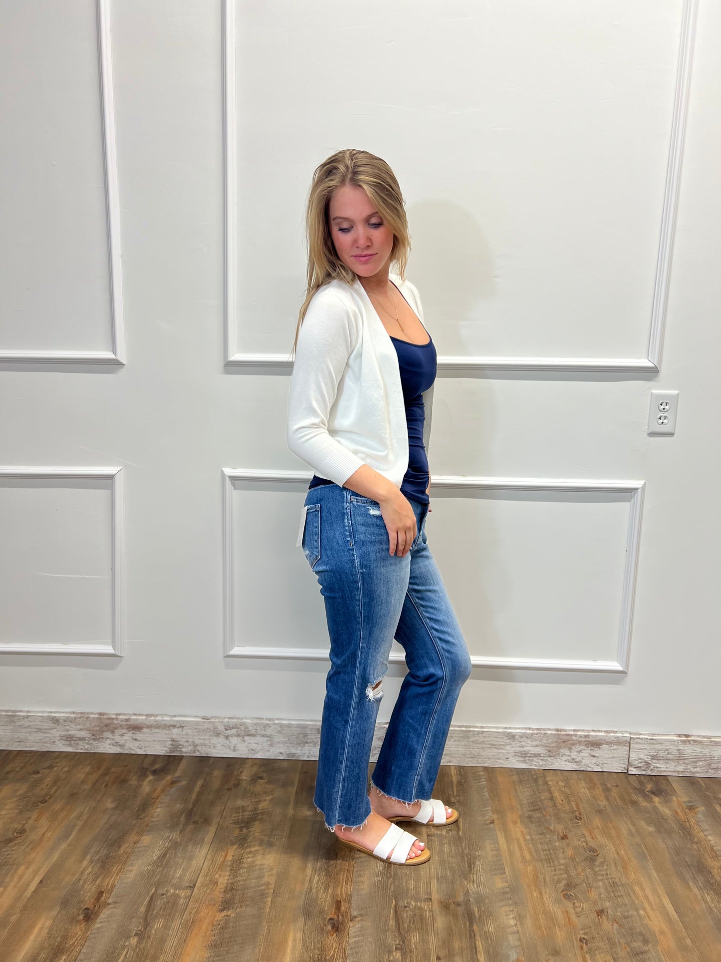 Woman wearing a white cardigan, blue top, and jeans standing in a room with a neutral wall.