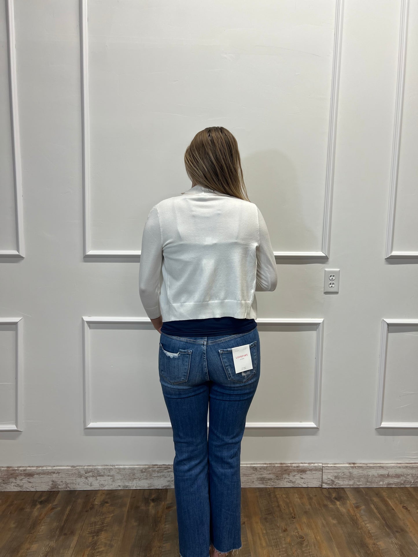 Person wearing a white jacket and blue jeans standing against a white paneled wall.