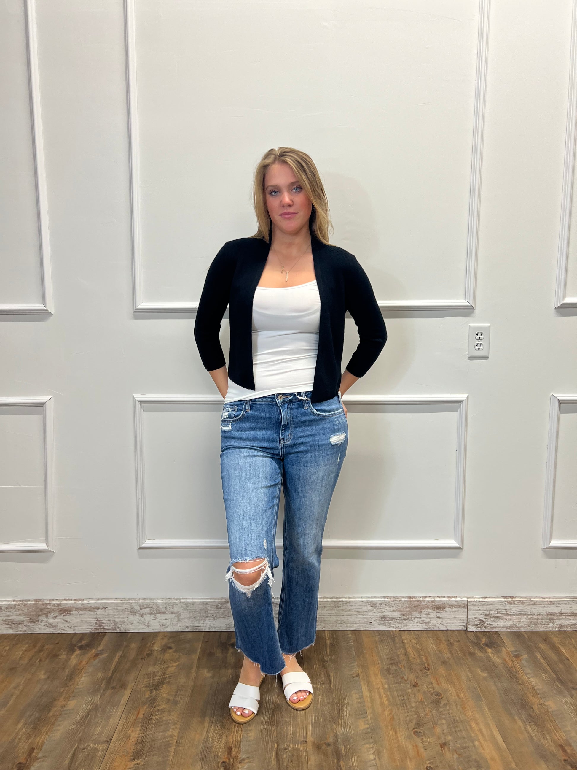 Person wearing a black cardigan, white top, and blue jeans standing in front of a white door.