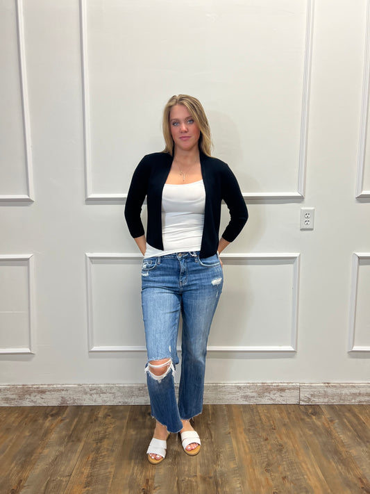 Person wearing a black cardigan, white top, and blue jeans standing in front of a white door.