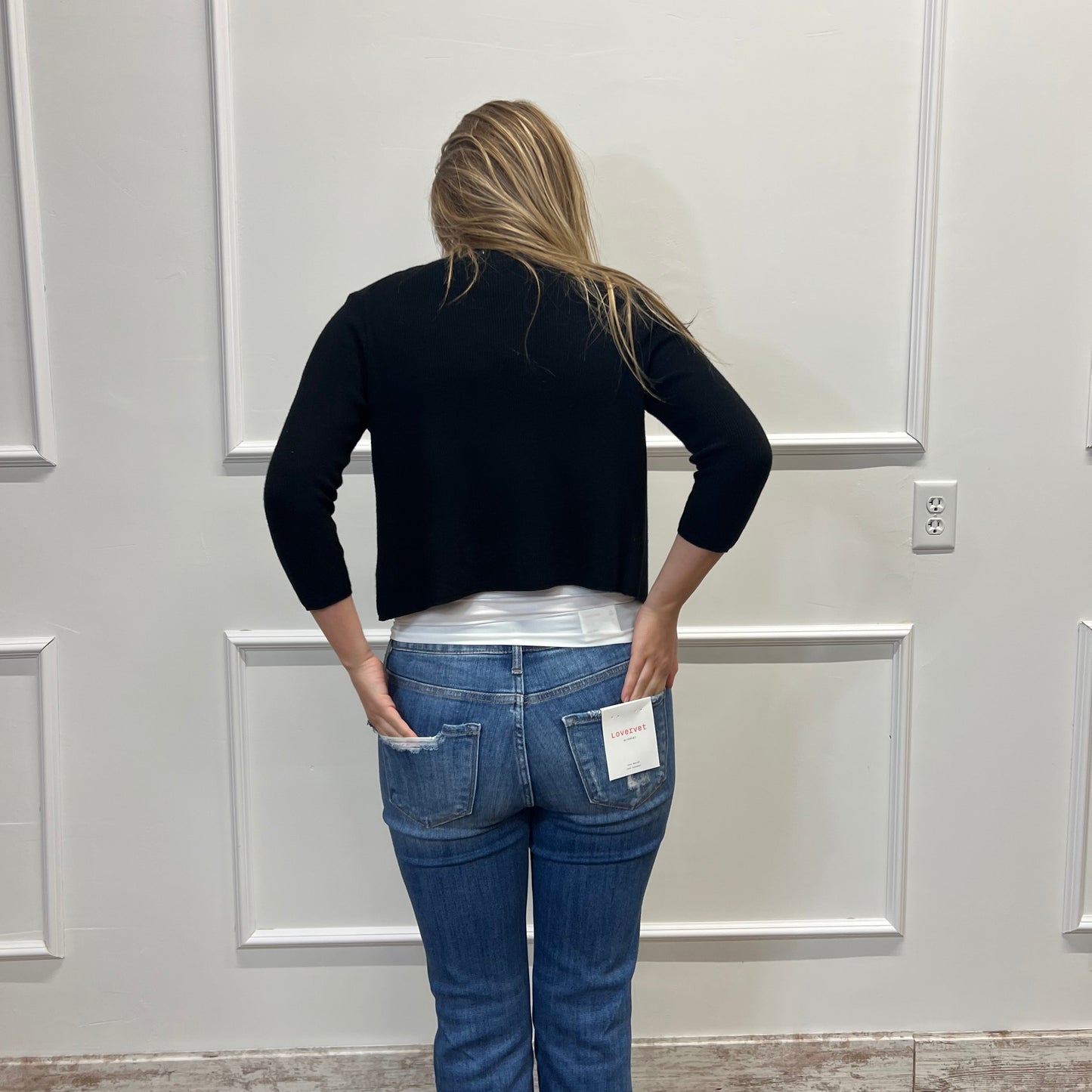 Person wearing a black top and blue jeans standing in front of a white door.