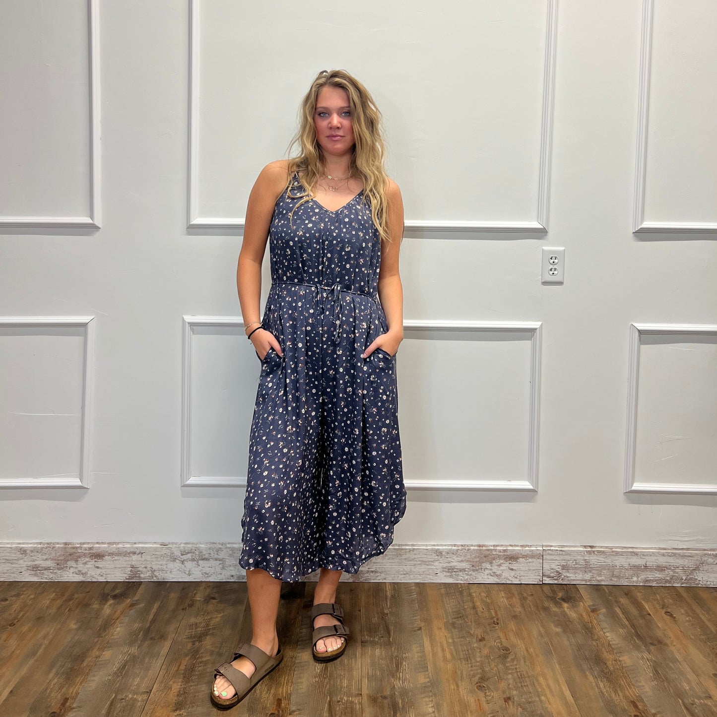 A woman modeling a sleeveless navy blue midi dress with a white floral pattern, standing in front of a white paneled door on a wood floor.