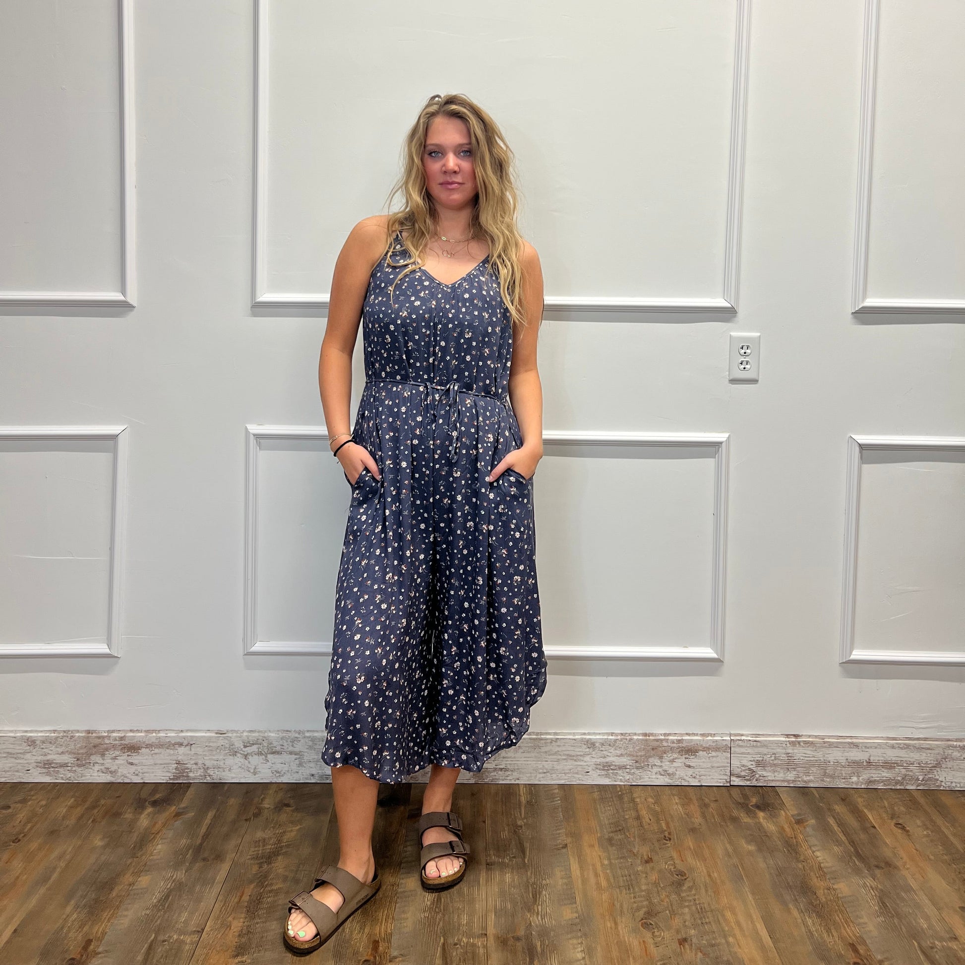 A woman modeling a sleeveless navy blue midi dress with a white floral pattern, standing in front of a white paneled door on a wood floor.