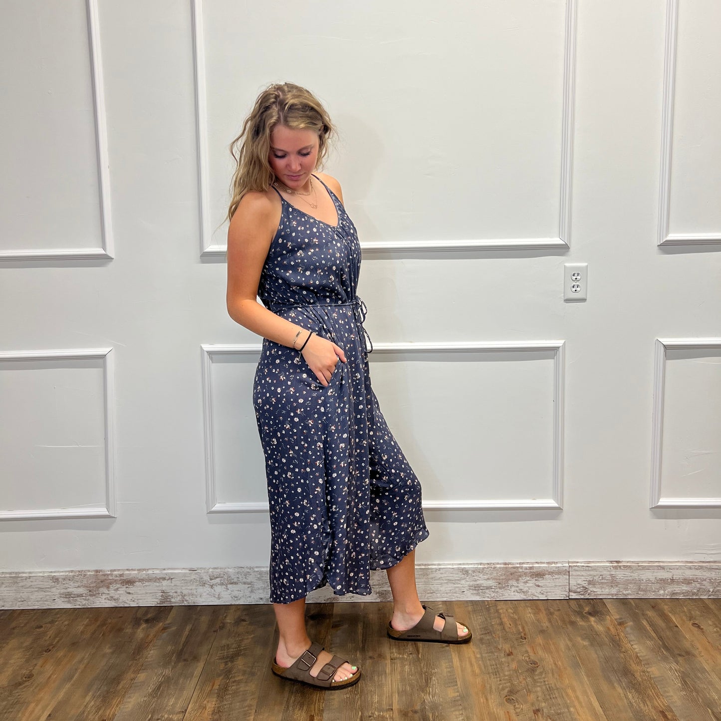 A woman wearing a navy blue sleeveless maxi dress with a white floral pattern, standing in front of a white paneled door on a wood floor.