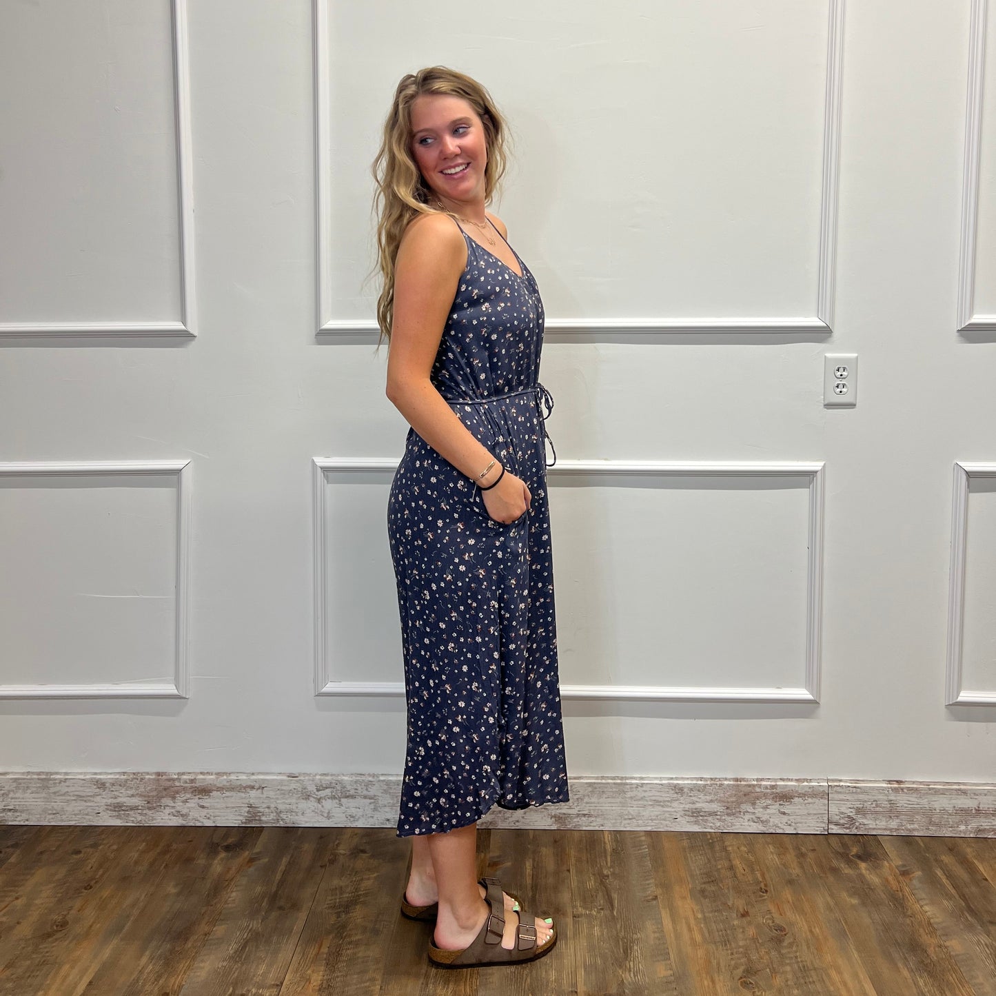 A woman wearing a navy blue sleeveless maxi dress with a white floral pattern, standing in front of a white paneled door.