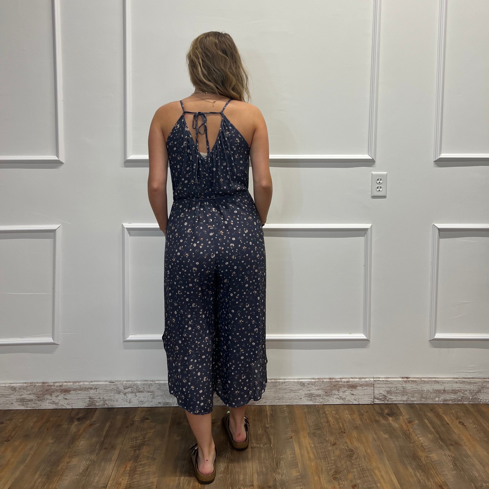 A woman viewed from behind wearing a navy blue and white micro-patterned halter jumpsuit, standing in front of a white paneled wall with a wood floor.