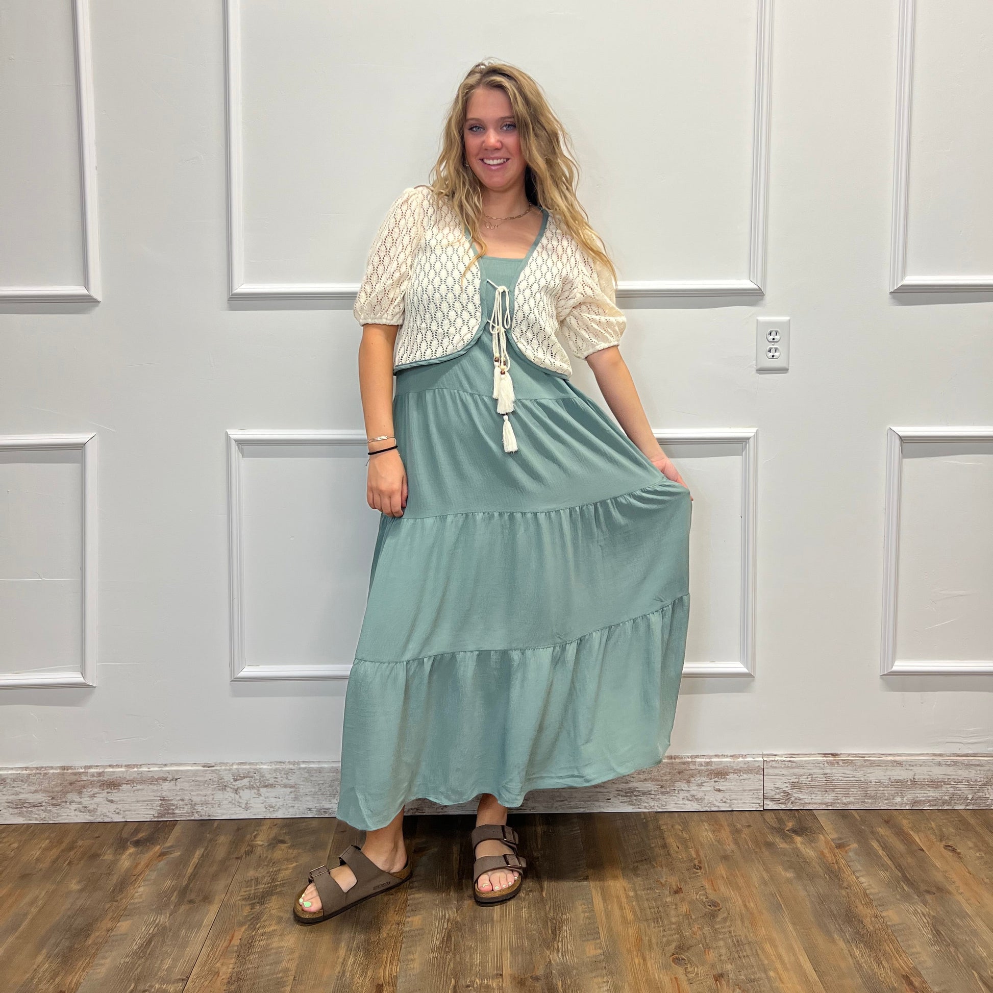 A woman wearing a sage green tiered maxi dress with a white textured short-sleeve top underneath, standing in front of a white paneled wall on a wood floor.