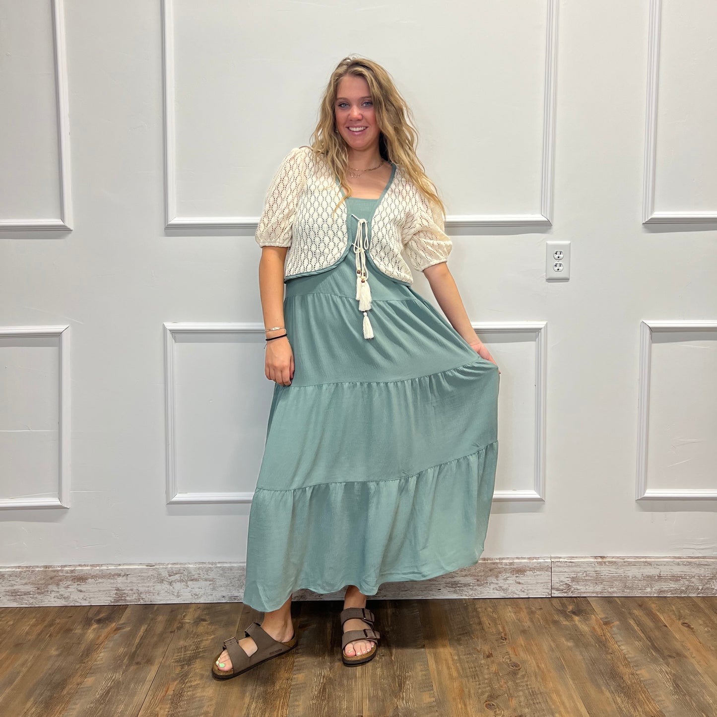 A woman wearing a sage green tiered maxi dress with a white textured short-sleeve top underneath, standing in front of a white paneled wall on a wood floor.