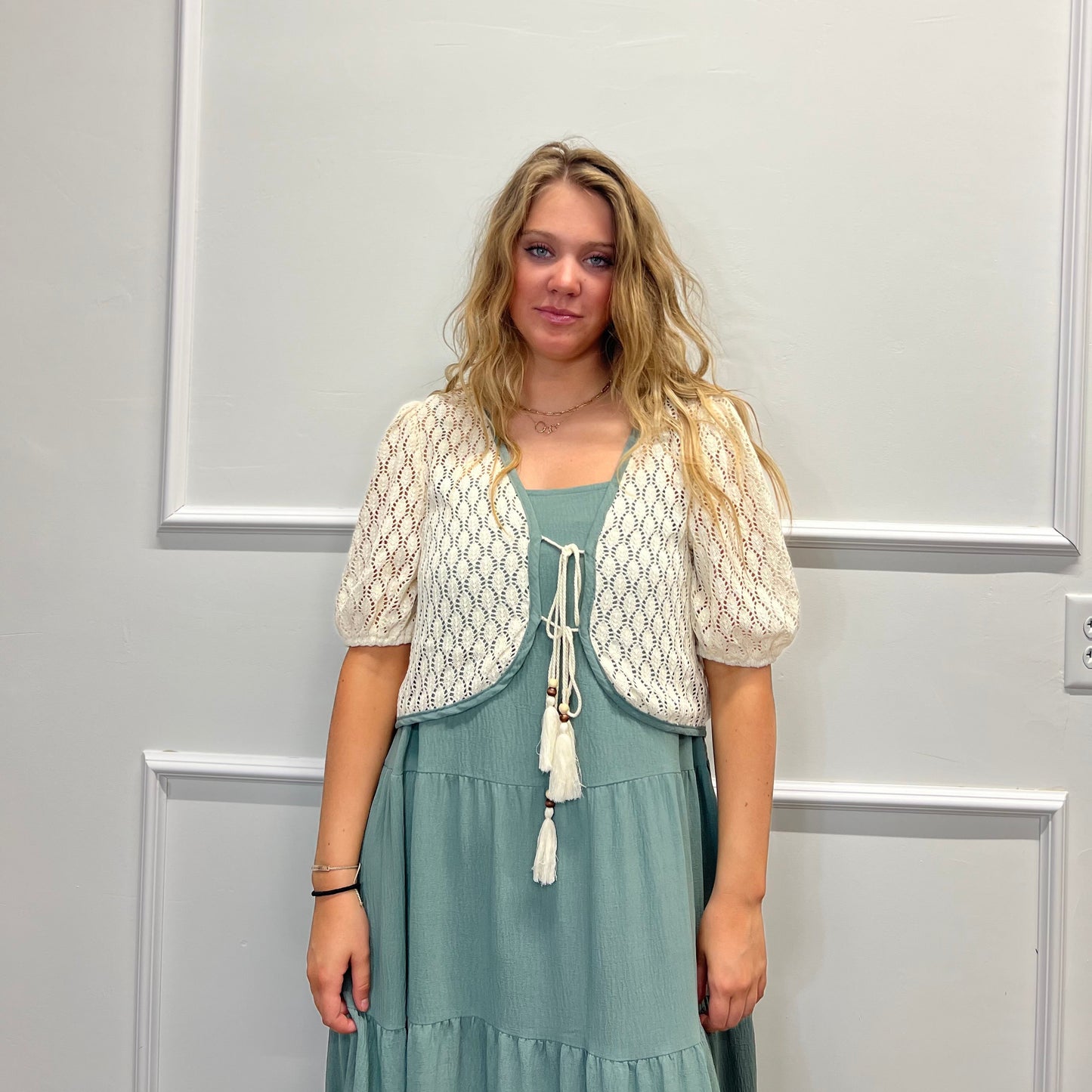 A woman stands against a white paneled wall wearing a cream-colored crochet shrug jacket over a dusty sage green tiered maxi dress.
