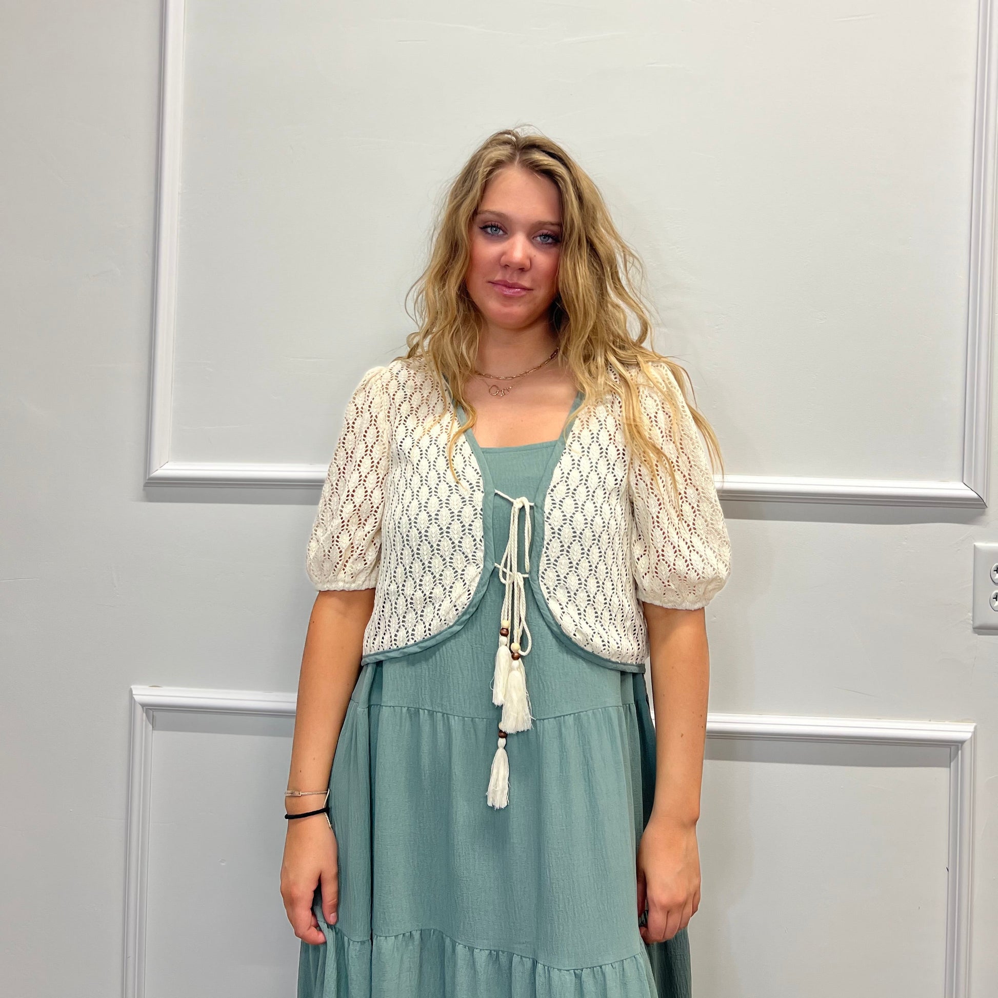 A woman stands against a white paneled wall wearing a cream-colored crochet shrug jacket over a dusty sage green tiered maxi dress.