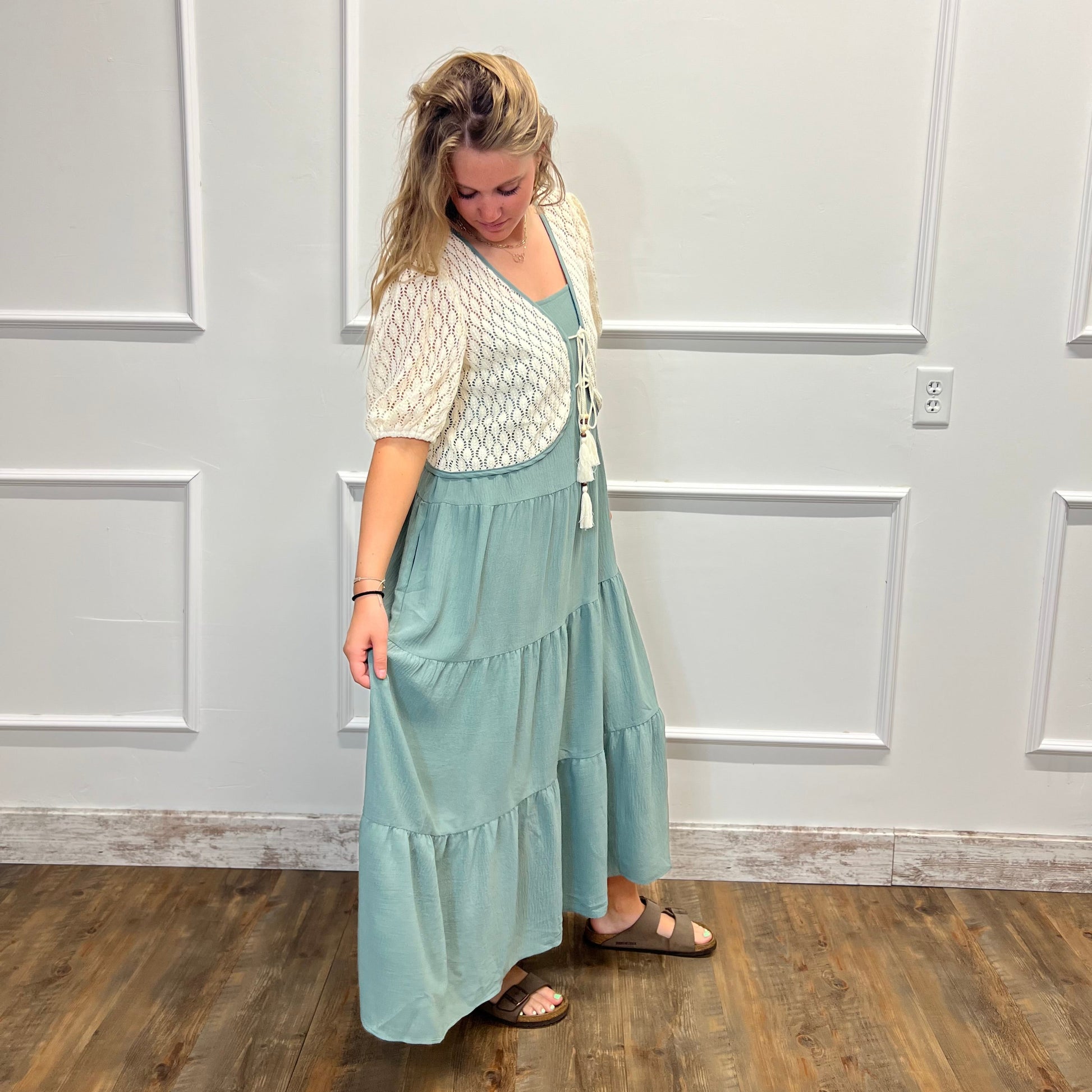 A woman modeling a two-piece outfit consisting of a light sage green tiered maxi skirt and a cream-colored textured short-sleeve cardigan, standing in front of a white paneled wall on a wooden floor.