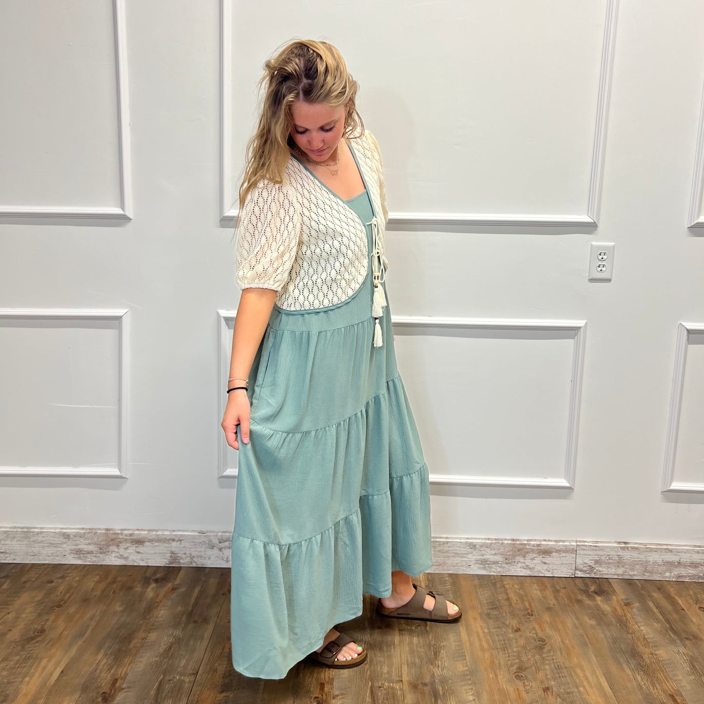 A woman modeling a two-piece outfit consisting of a light sage green tiered maxi skirt and a cream-colored textured short-sleeve cardigan, standing in front of a white paneled wall on a wooden floor.