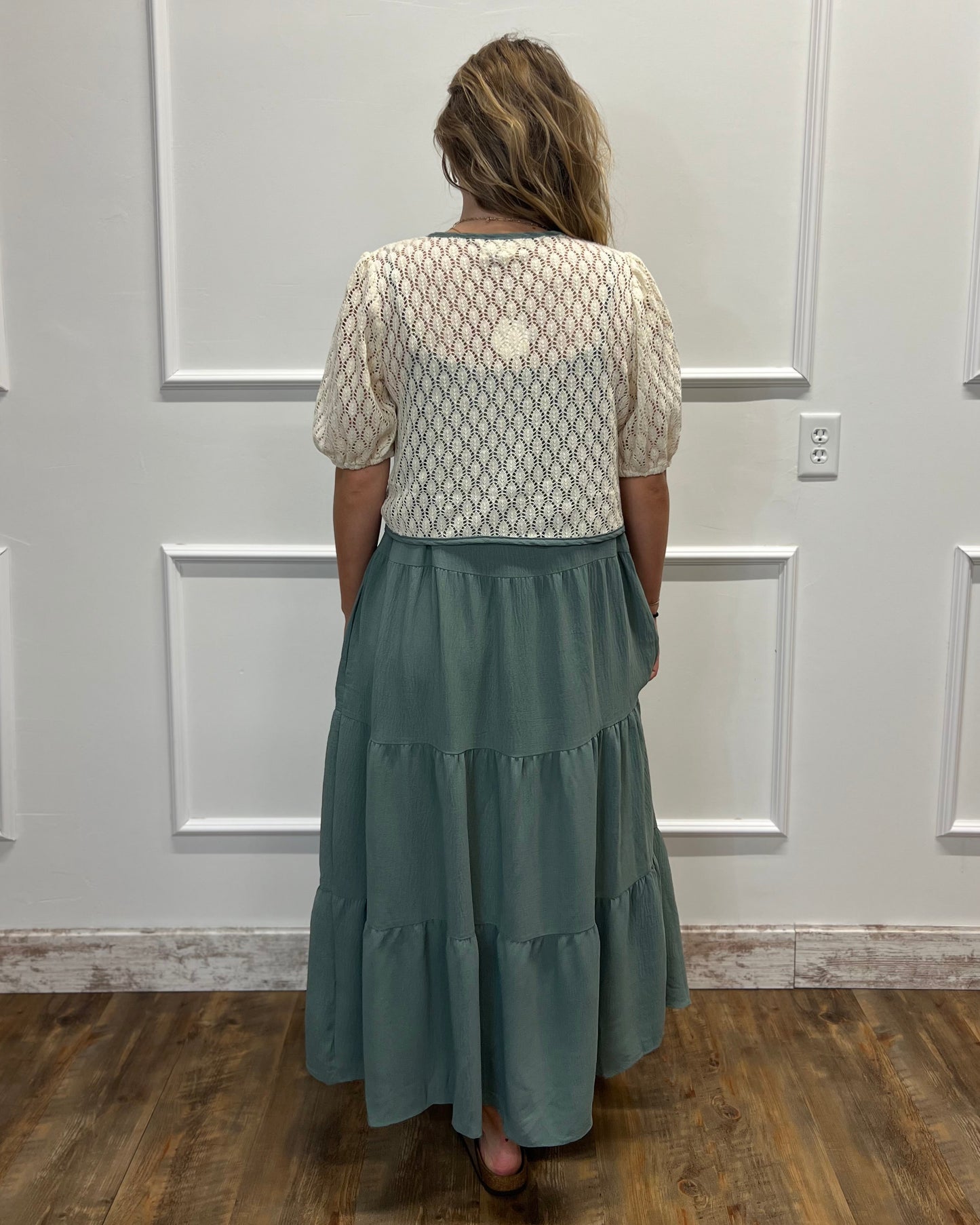A woman viewed from behind wearing a cream-colored textured short-sleeve top and a tiered sage green maxi skirt, standing in front of a white paneled wall on a wooden floor.