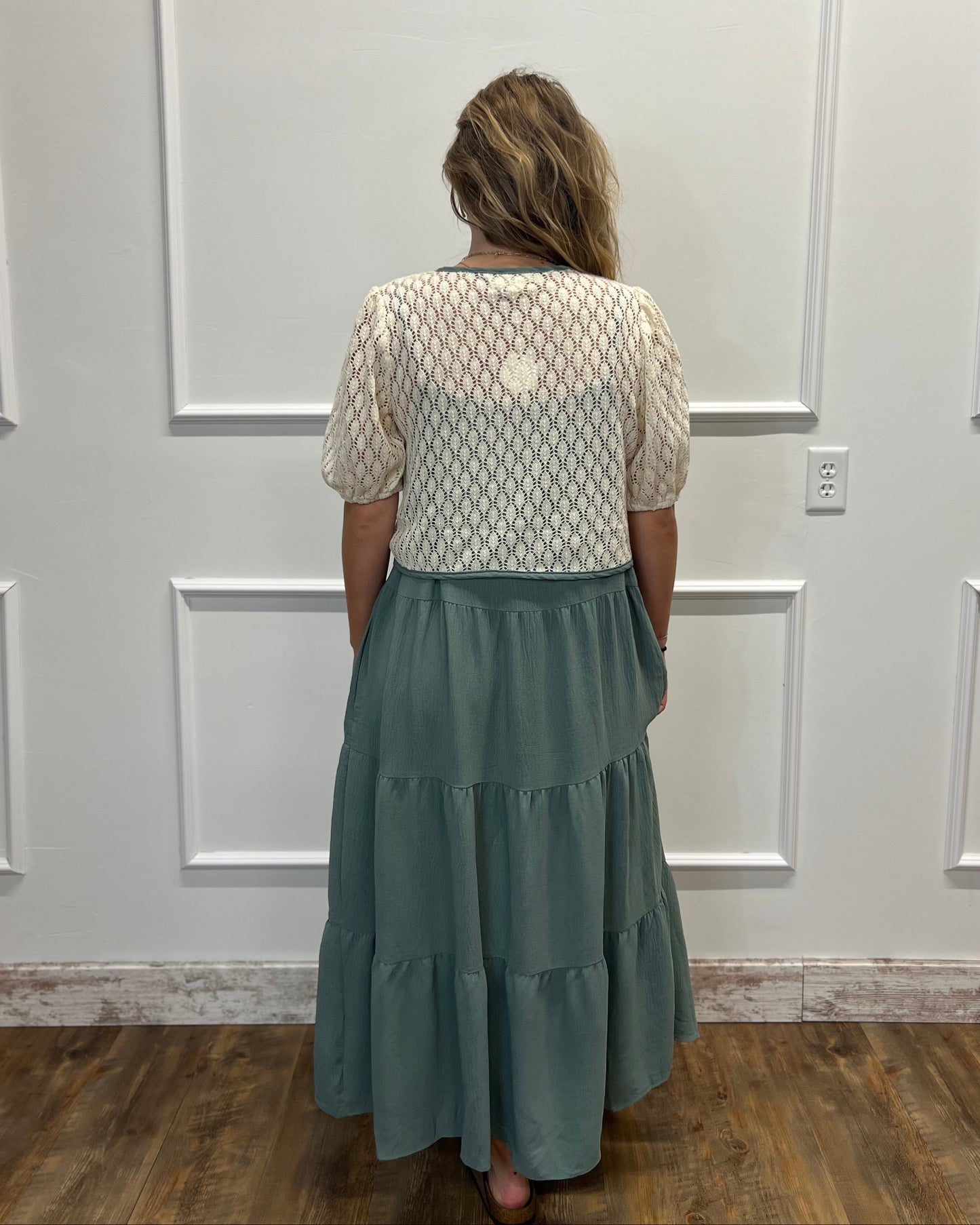 A woman viewed from behind wearing a cream-colored textured short-sleeve top and a tiered sage green maxi skirt, standing in front of a white paneled wall on a wooden floor.
