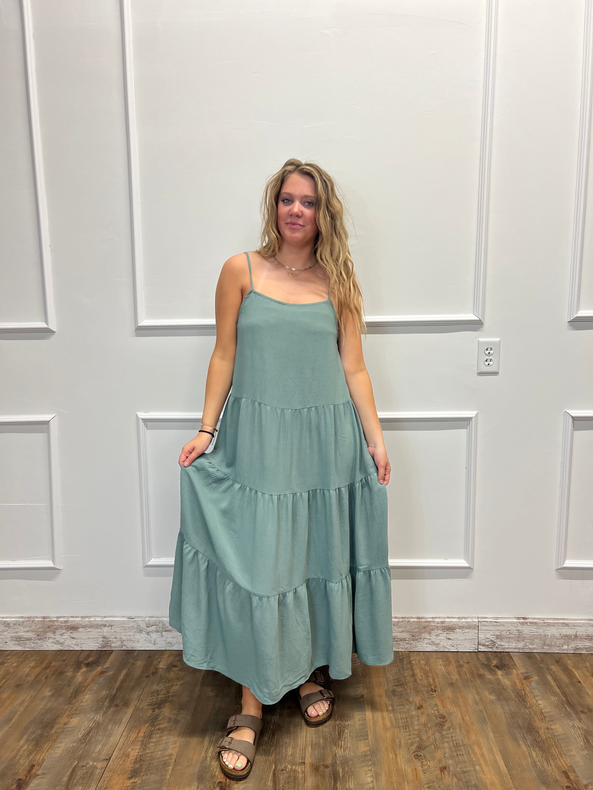 A woman wearing a sleeveless tiered maxi dress in sage green, standing in front of a white paneled door on a wood floor.