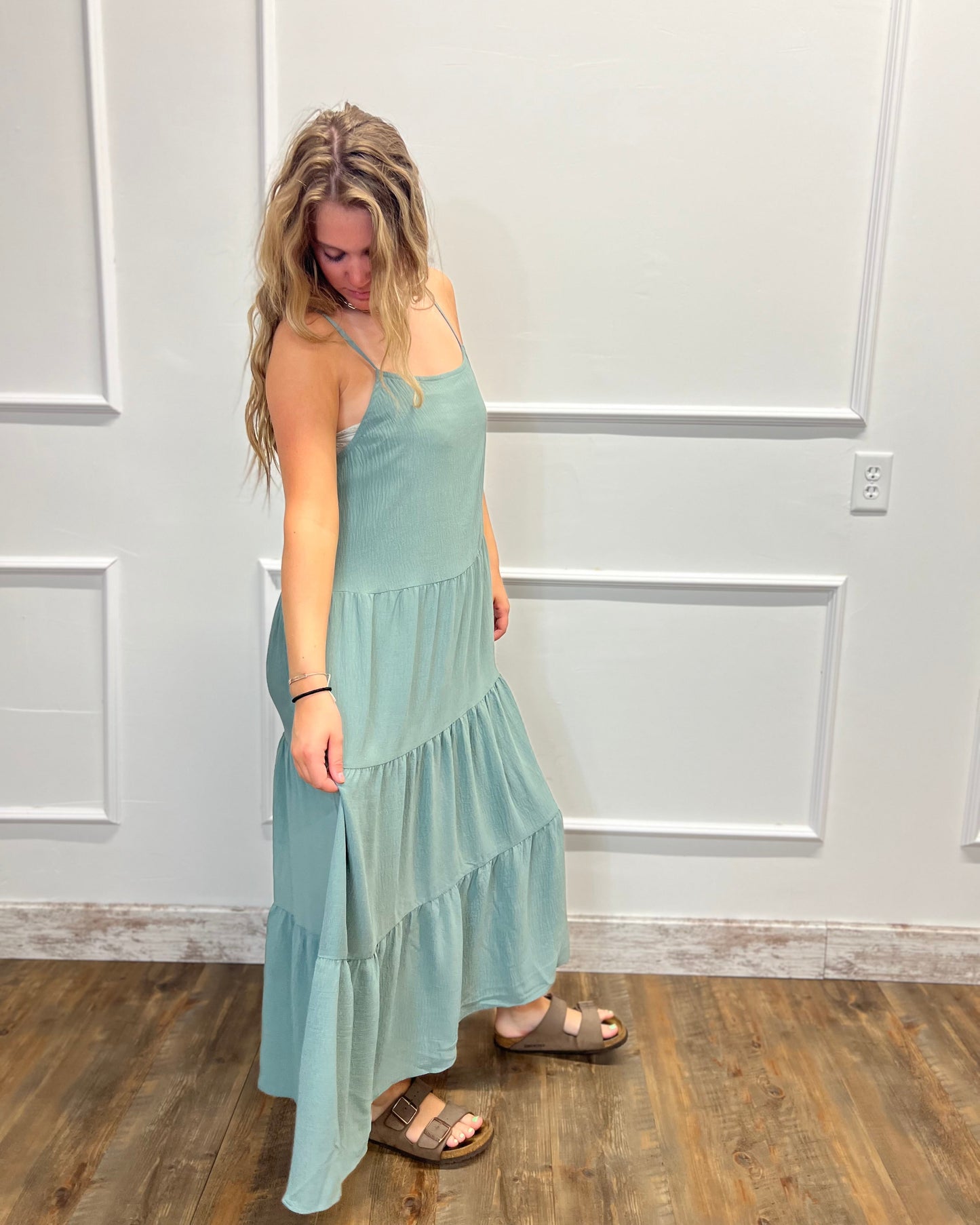 A woman modeling a light blue-green tiered maxi dress with spaghetti straps, paired with brown slide sandals, standing in front of a white paneled wall.