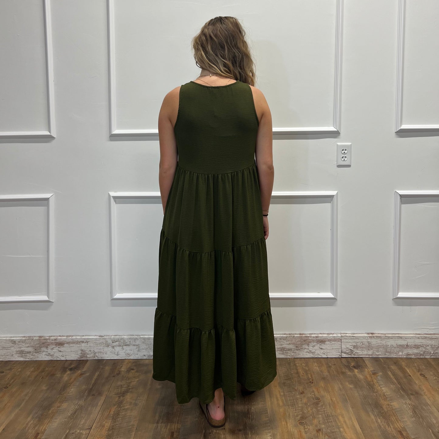 A woman viewed from behind wearing a sleeveless olive green tiered maxi dress, standing on a wood floor against a white paneled wall.