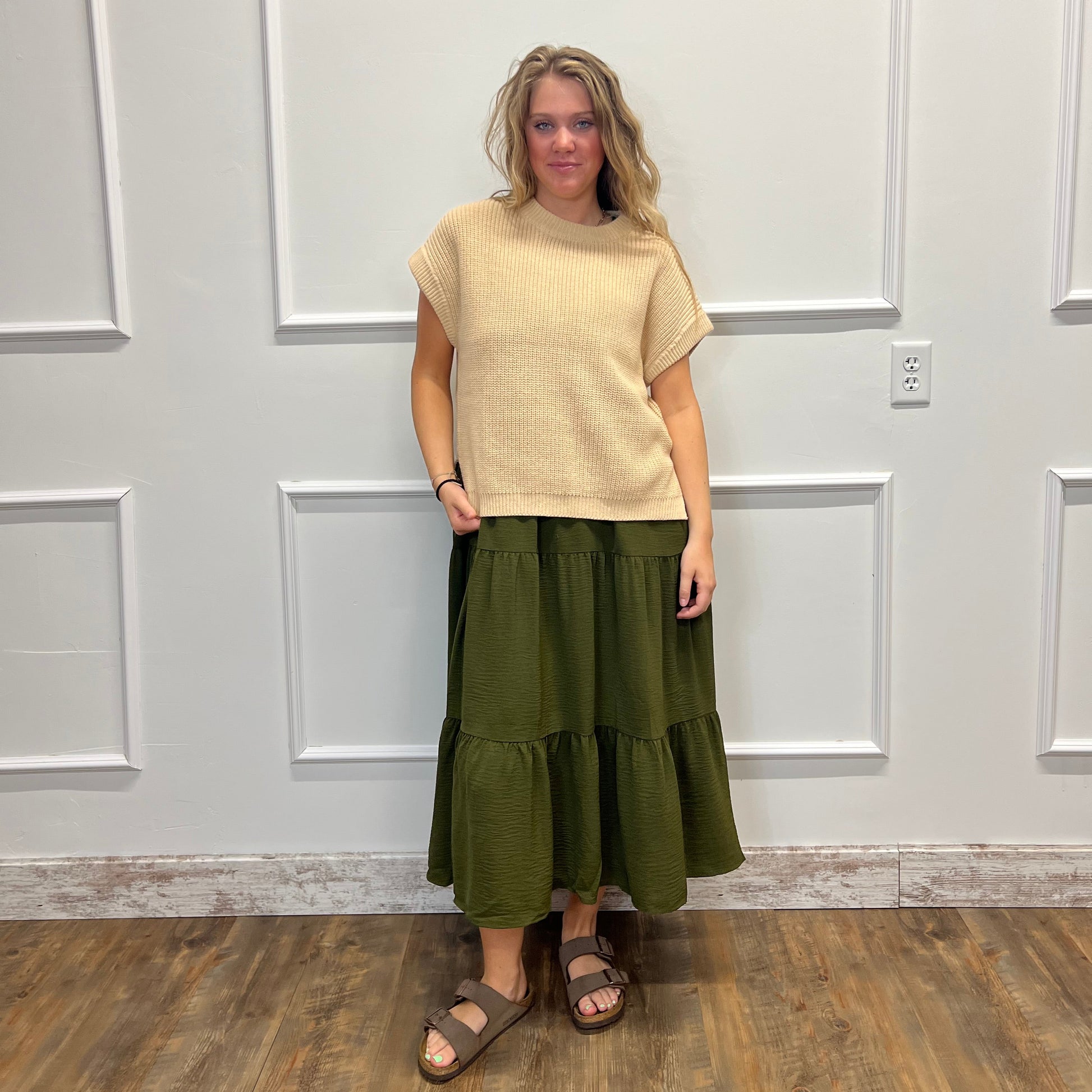 A woman wearing a beige short-sleeve knit top and an olive green tiered skirt, standing in front of a white paneled door on a wood floor.