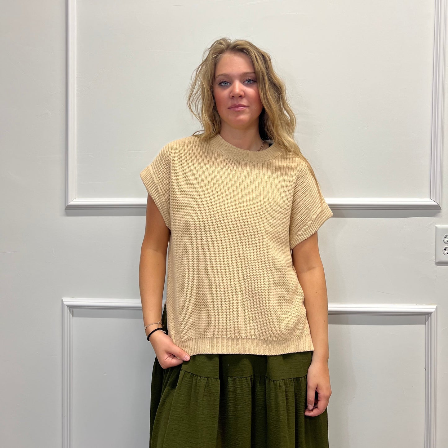 A woman stands against a white paneled wall wearing a beige short-sleeve textured knit top and an olive green skirt.