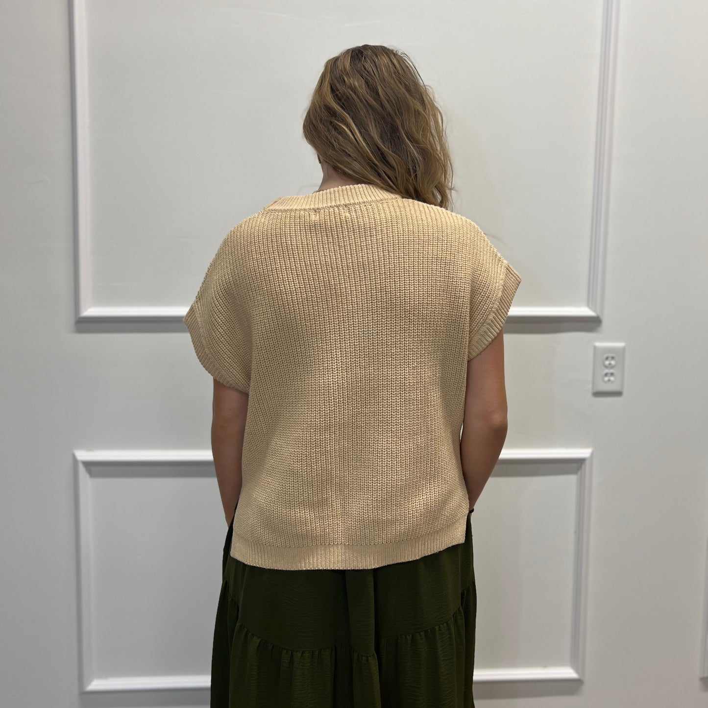 A woman viewed from behind wearing a beige short-sleeve ribbed knit top and an olive green skirt, standing in front of a white paneled wall.