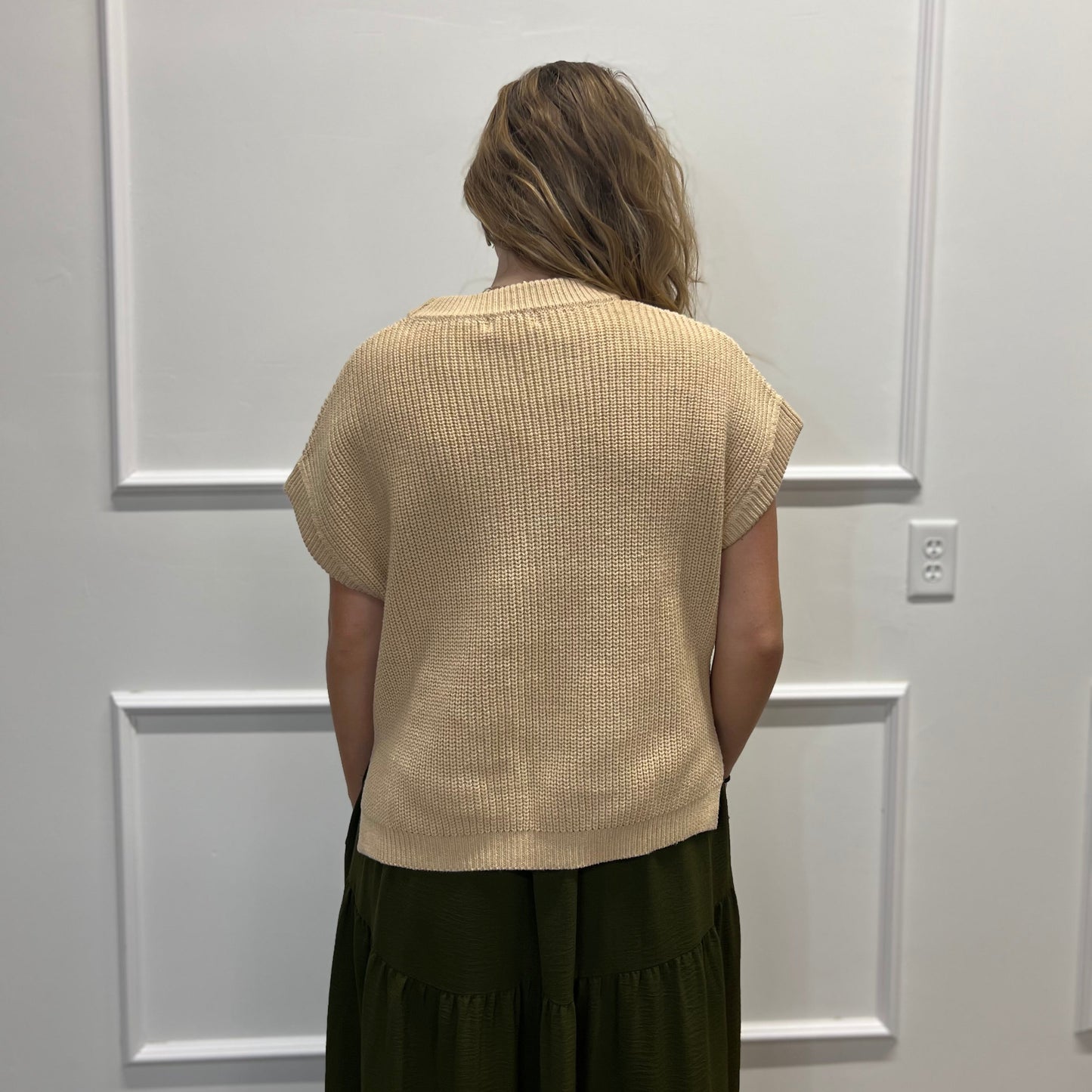 A woman viewed from behind wearing a beige short-sleeve ribbed knit top and a dark olive green skirt, standing against a white paneled wall.