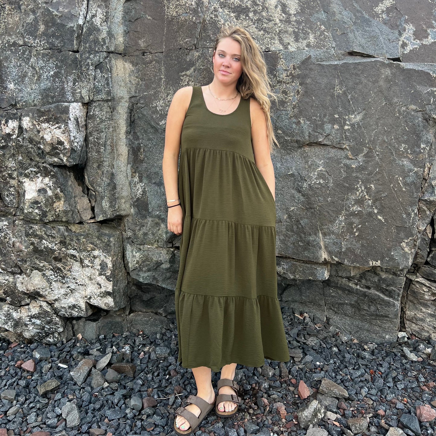 A woman wearing an olive green sleeveless tiered maxi dress and brown sandals, standing outdoors in front of a large rock wall on a bed of dark pebbles.