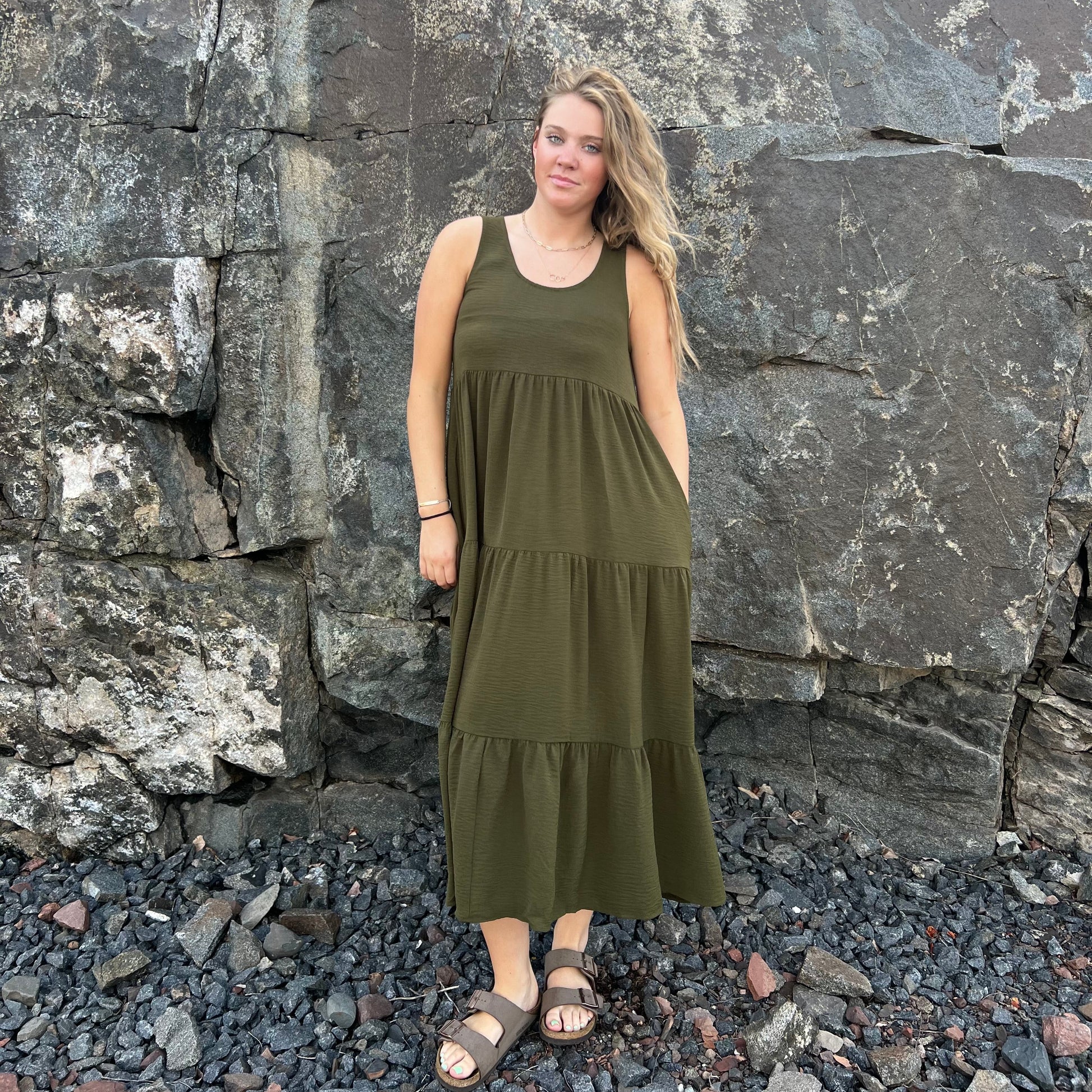 A woman wearing an olive green sleeveless tiered maxi dress and brown sandals, standing outdoors in front of a large rock wall on a bed of dark pebbles.