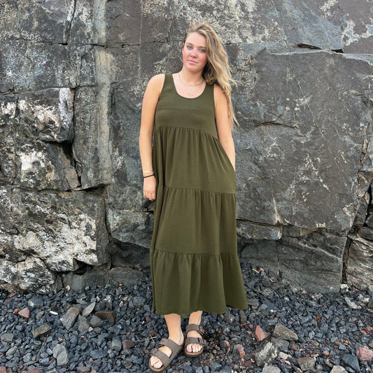 A woman wearing an olive green sleeveless tiered maxi dress and brown sandals, standing outdoors in front of a large rock wall on a bed of dark pebbles.