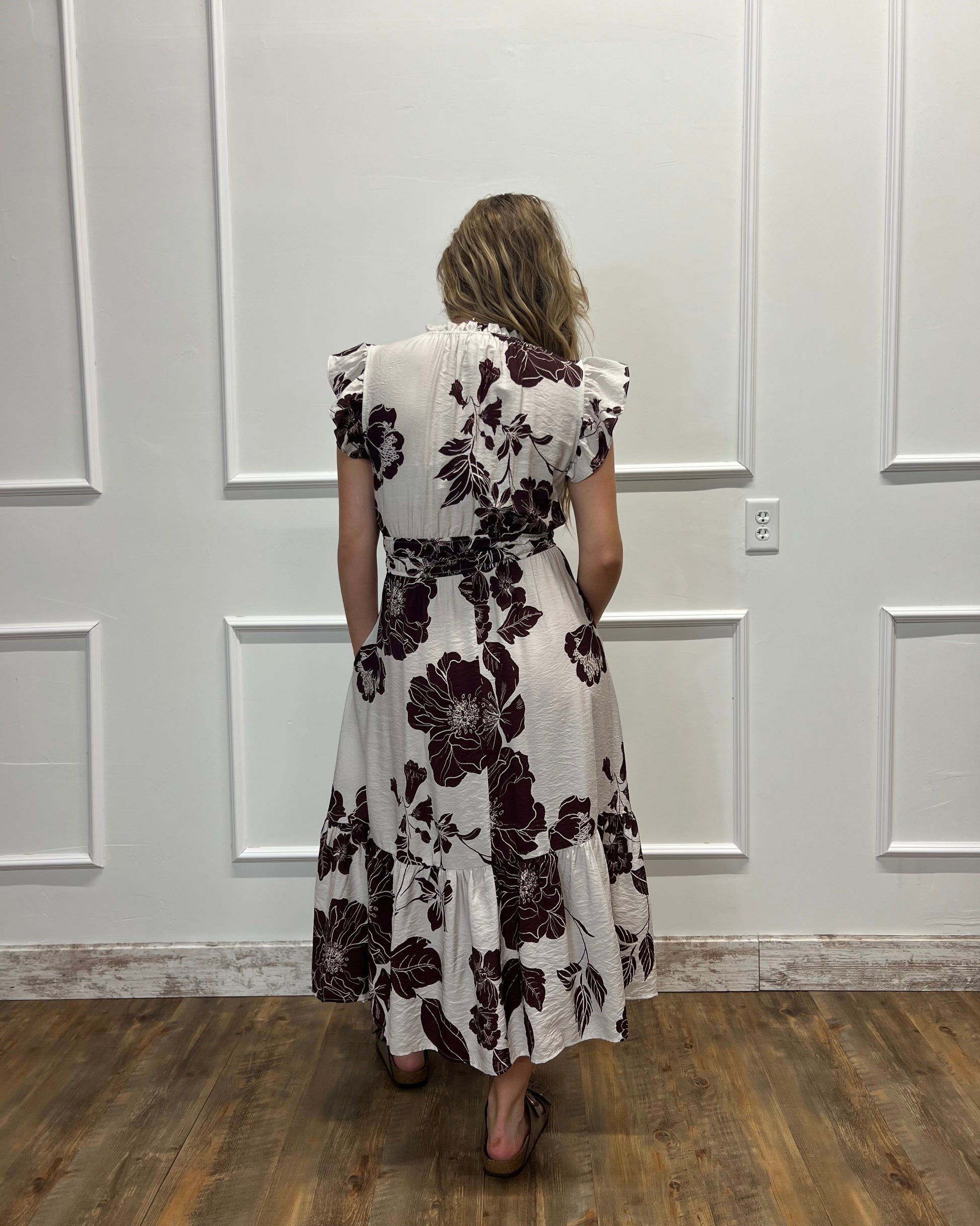 A woman viewed from behind wearing a white midi dress with a dark purple floral pattern, standing in front of a white paneled wall on a wooden floor.