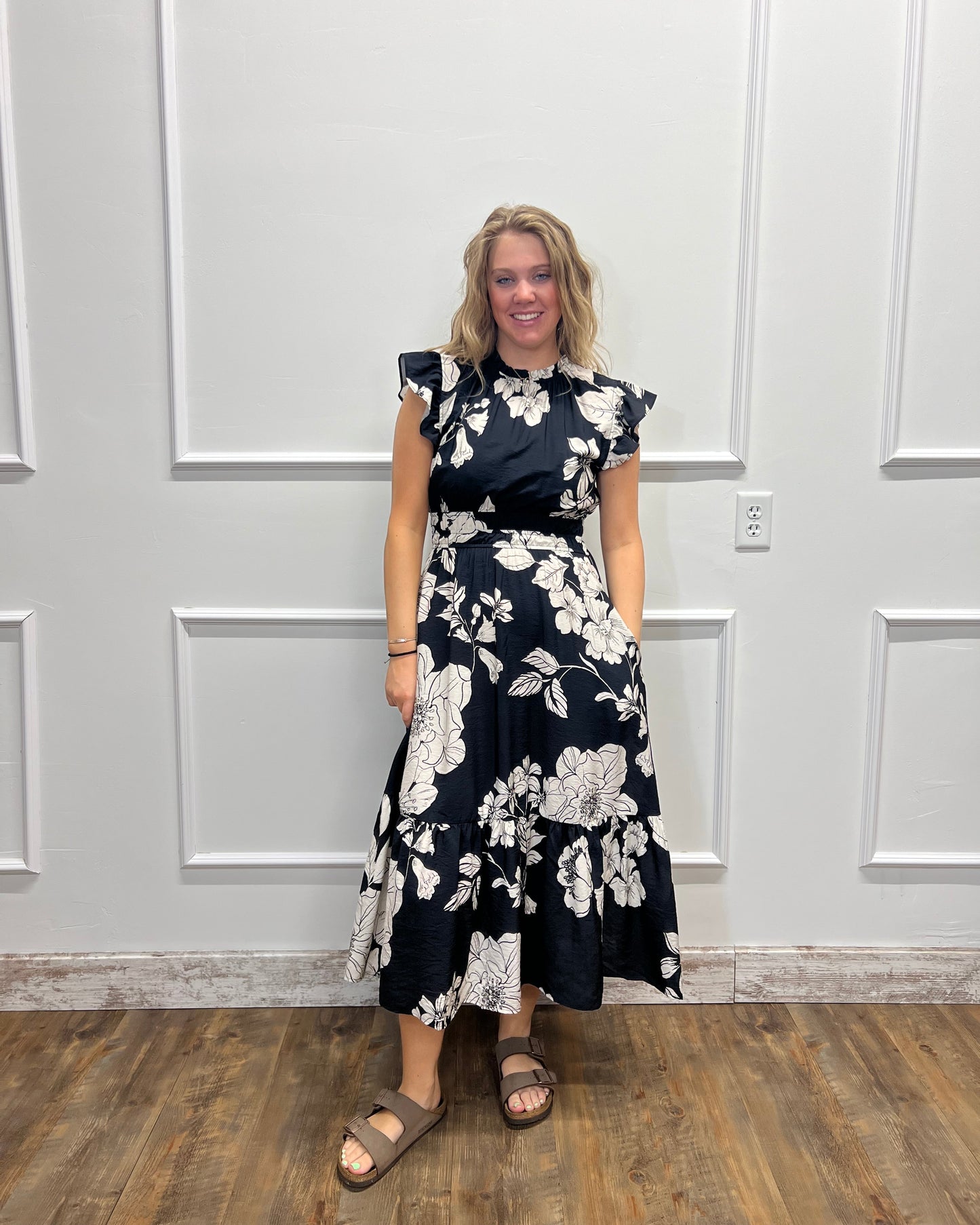 A woman wearing a navy blue and white floral print dress, standing in front of a white paneled wall with a wood floor visible on the right.