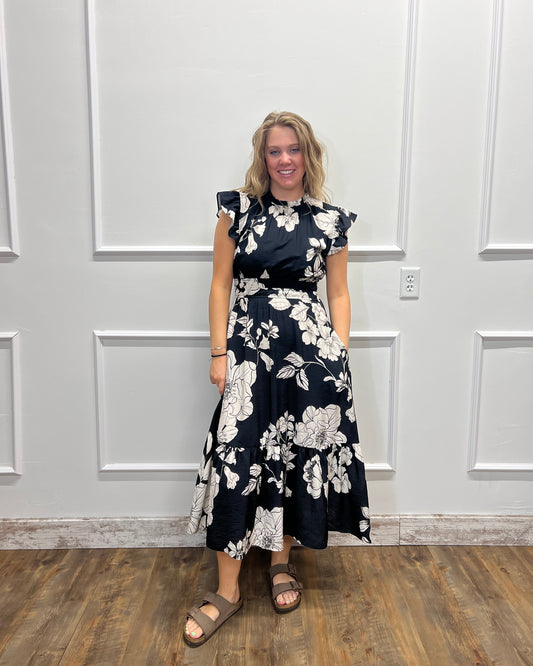 A woman wearing a navy blue and white floral print dress, standing in front of a white paneled wall with a wood floor visible on the right.