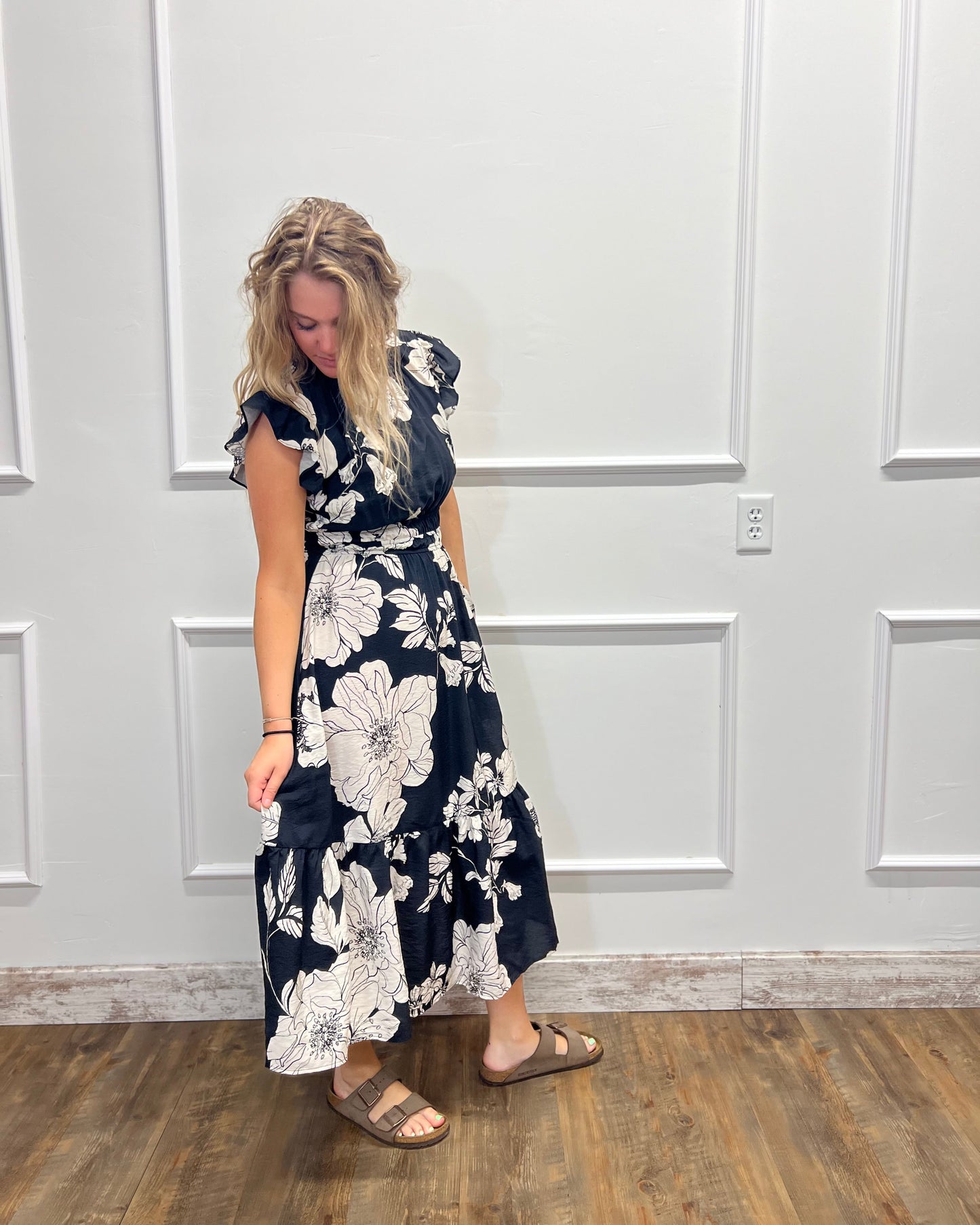 A woman wearing a navy blue midi dress with a large white floral print, standing in front of a white paneled wall on a wood floor.
