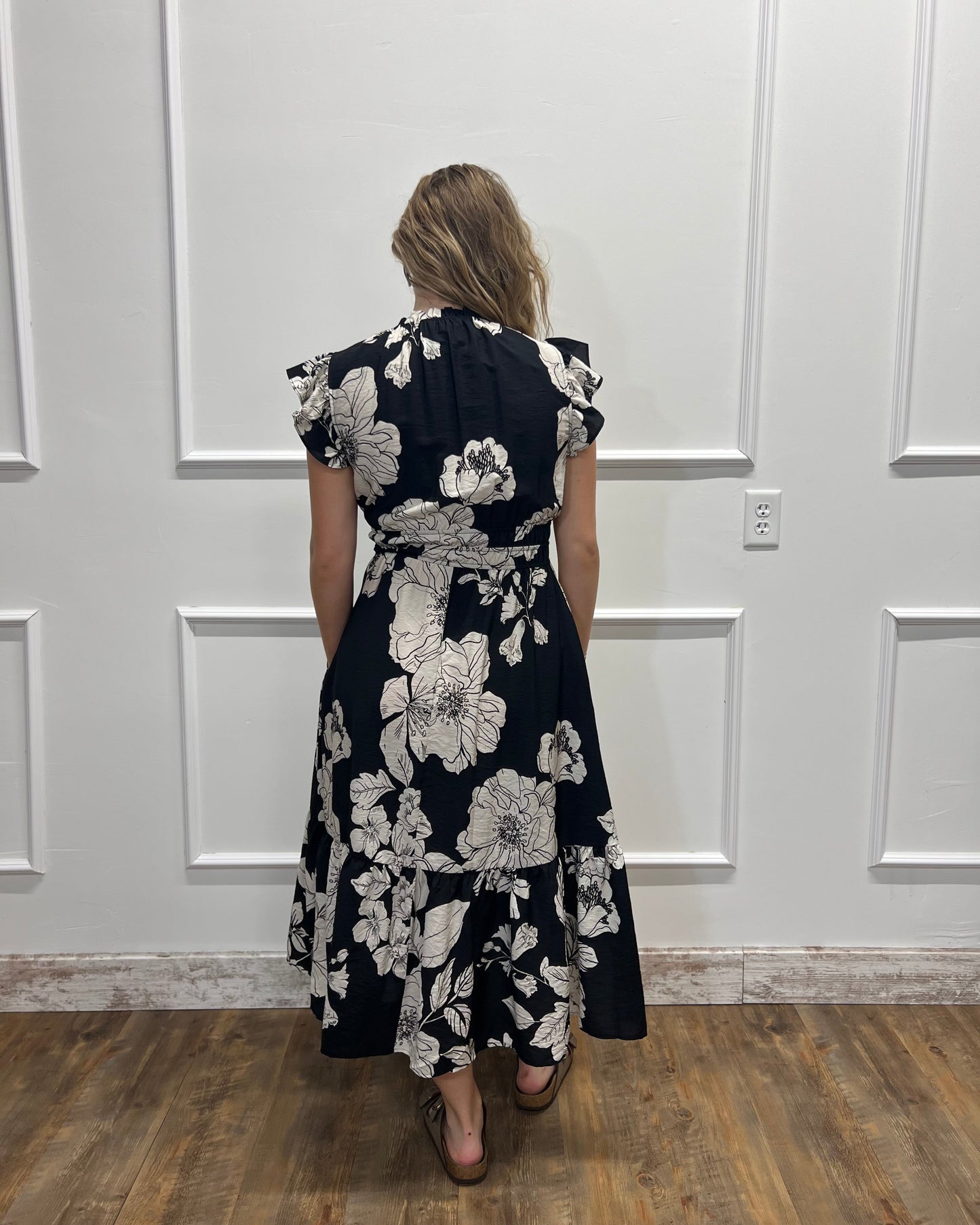 A woman viewed from behind wearing a black midi dress with a large white floral print, standing on a wood floor against a white paneled wall.