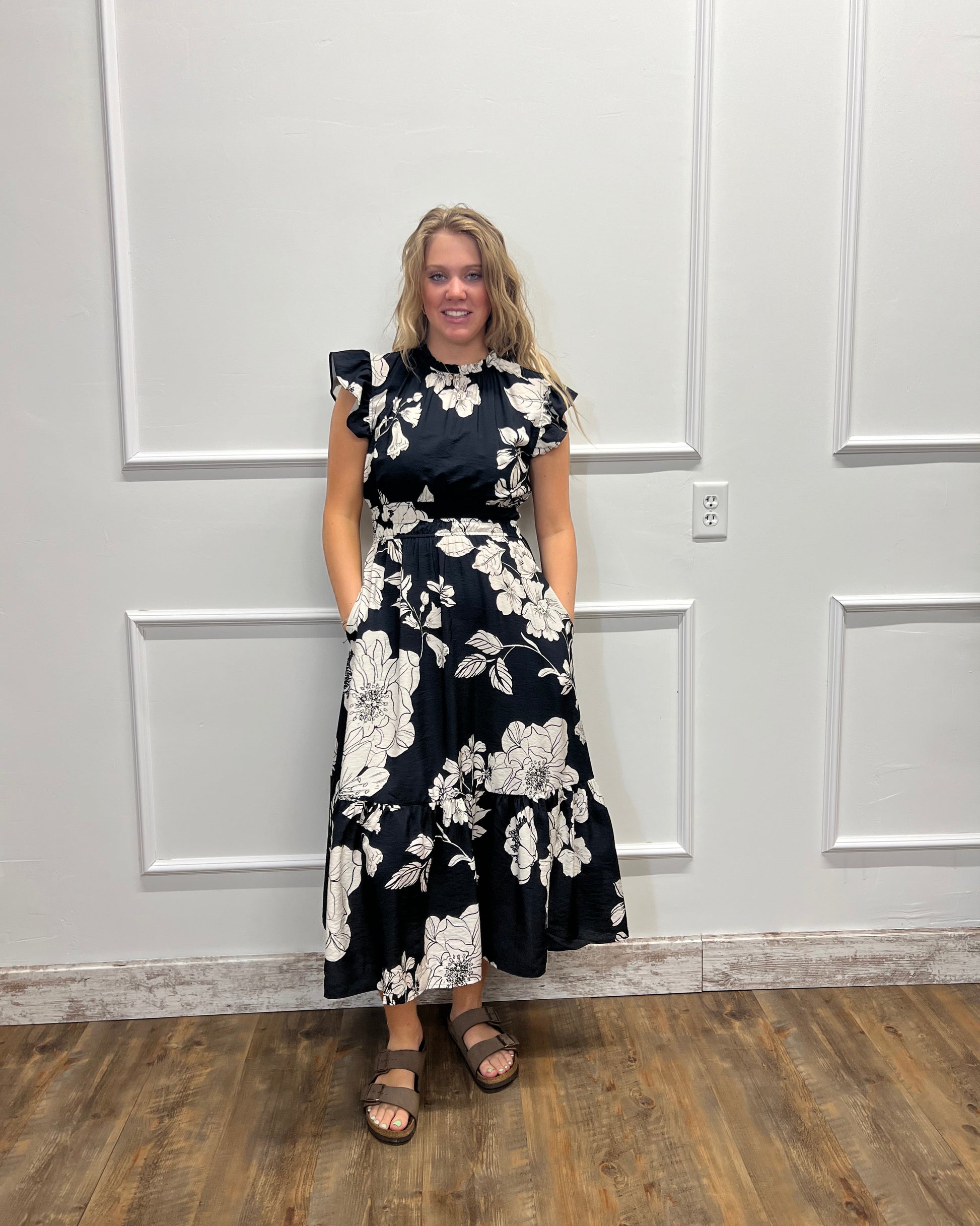 A woman wearing a black short-sleeve dress with a large white floral pattern, standing in front of a white paneled wall on a wood floor.