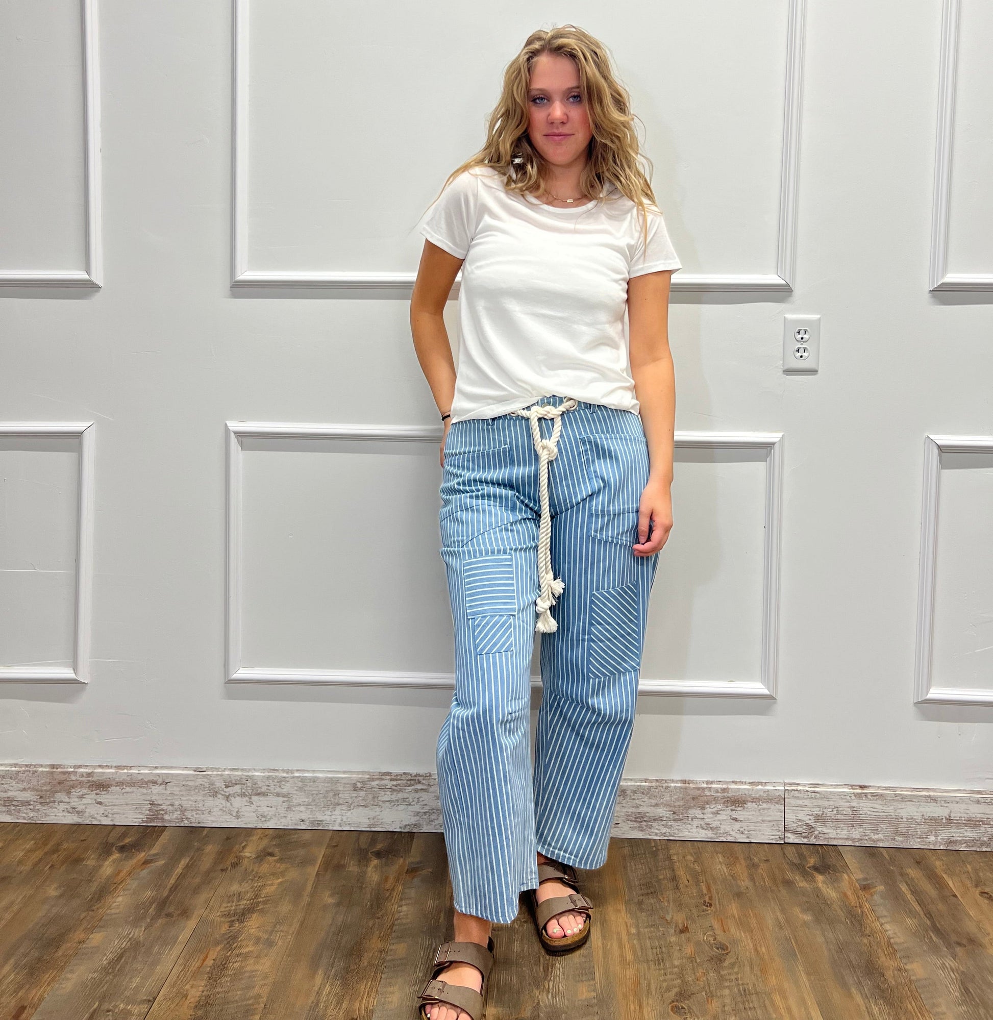 A woman wearing a white short-sleeve t-shirt and light blue pinstriped wide-leg pants with a white rope belt, standing in front of a white paneled wall.