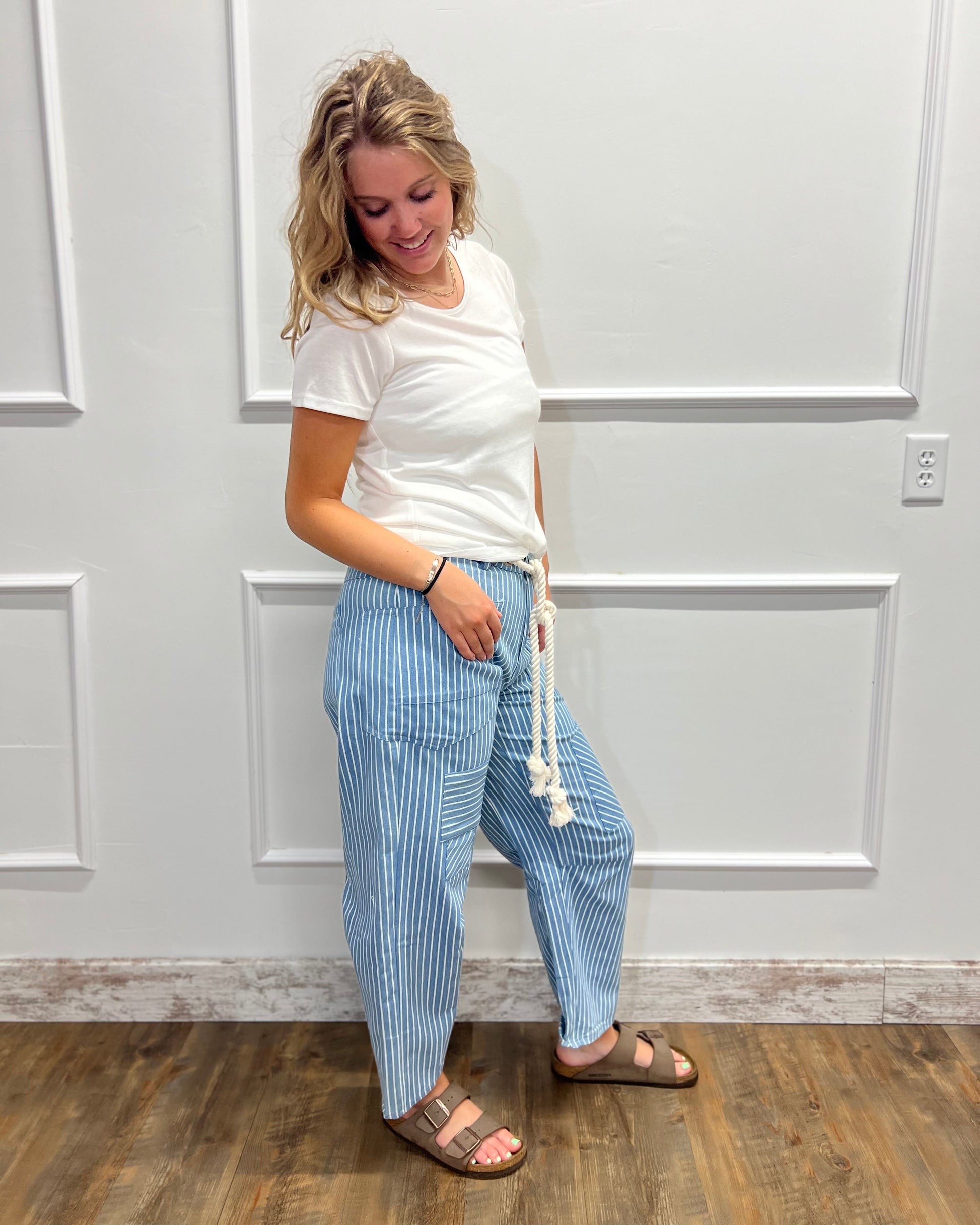 A woman modeling a pair of light blue and white vertically striped drawstring pants, paired with a white t-shirt and brown sandals, standing against a white paneled wall.
