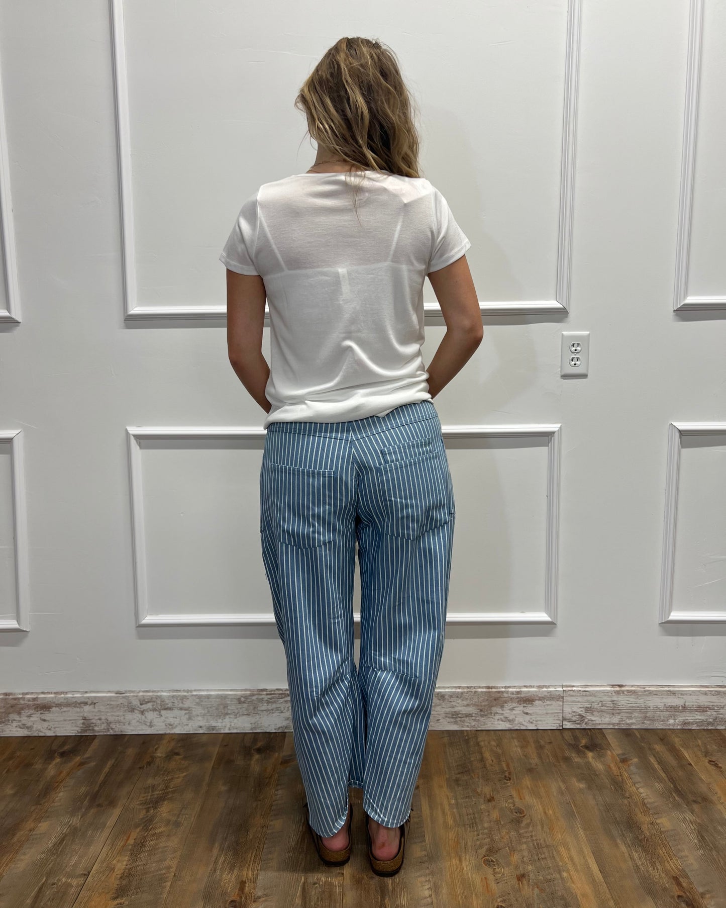 A woman viewed from behind wearing a white short-sleeve top and blue-and-white vertically striped trousers, standing on a wood floor against a white paneled wall.