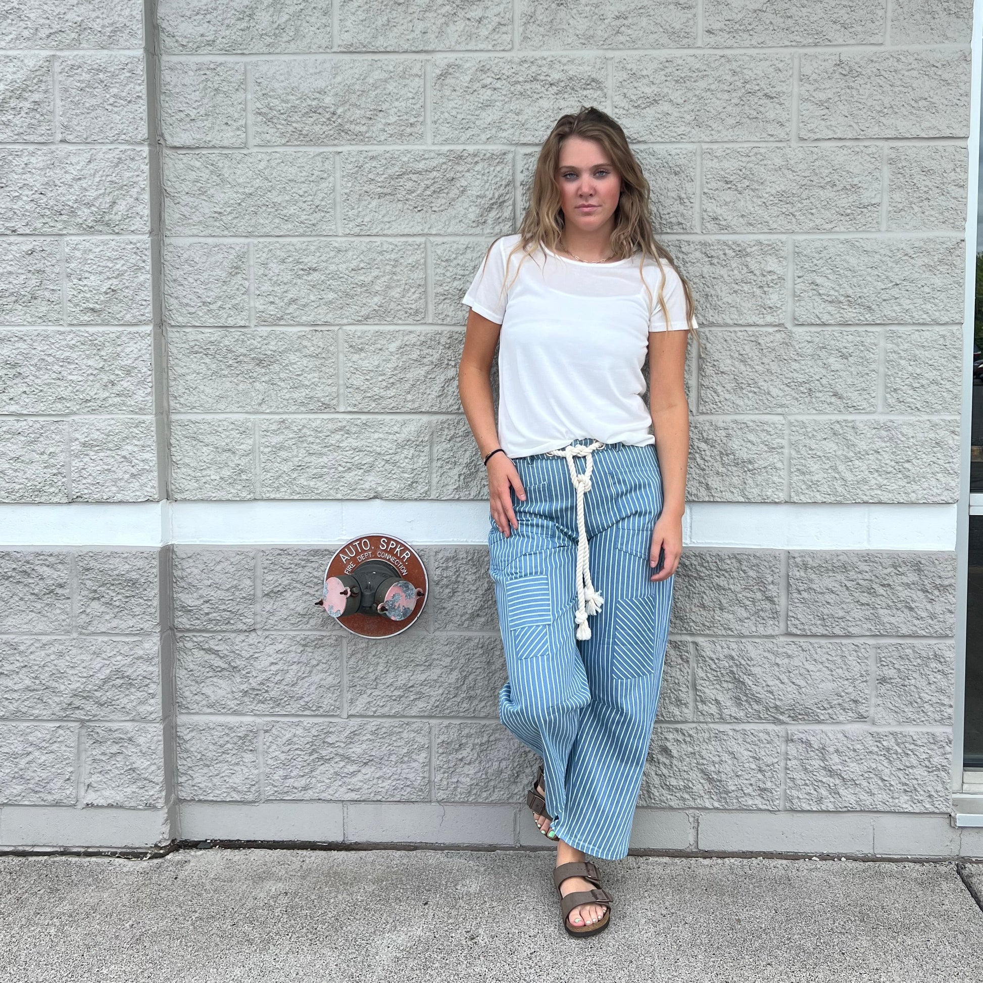 A woman wearing a white t-shirt and light blue striped wide-leg pants with a white drawstring, standing outdoors against a gray stone wall.