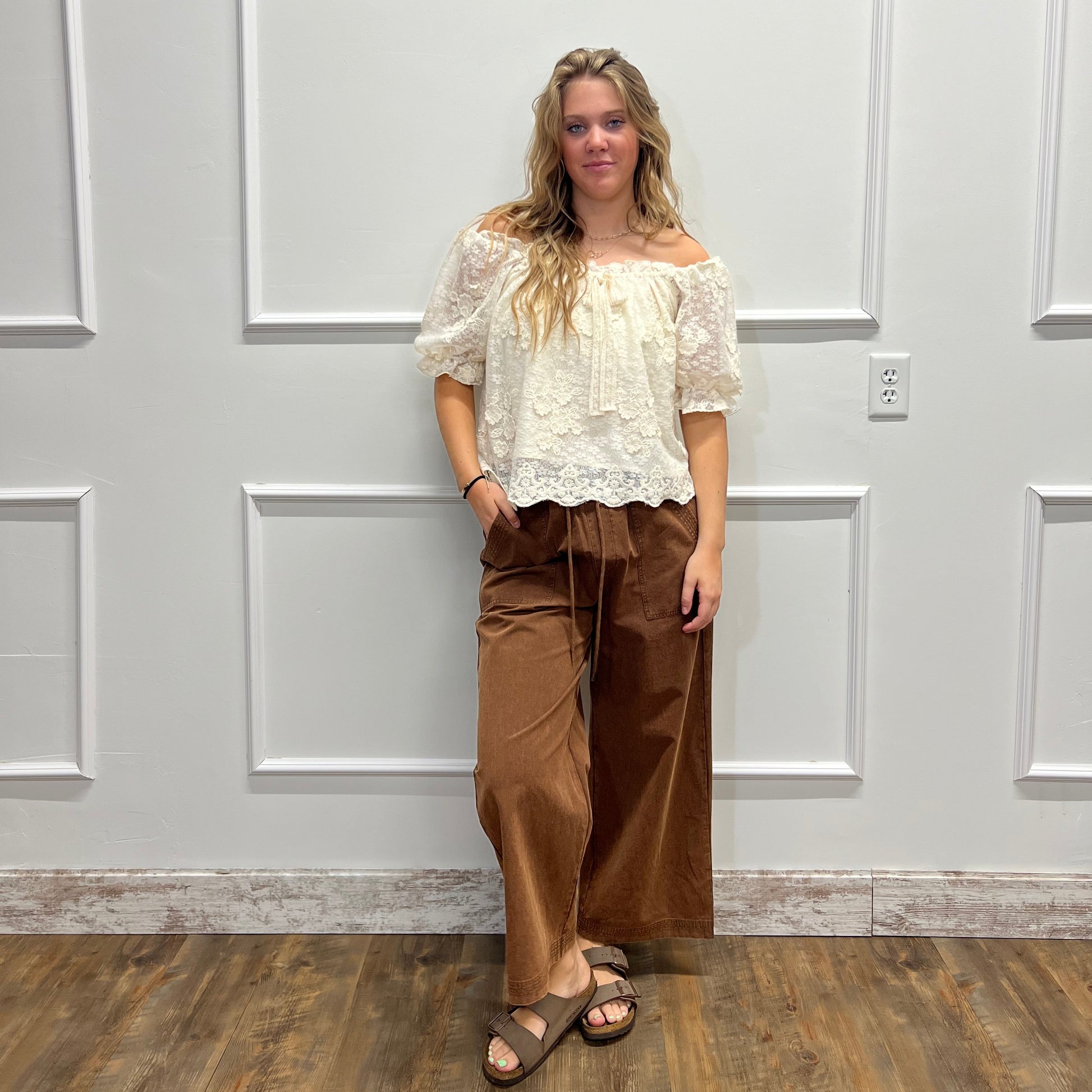 A woman modeling a two-piece outfit consisting of a cream lace short-sleeve top and brown wide-leg pants, standing in front of a white paneled door.